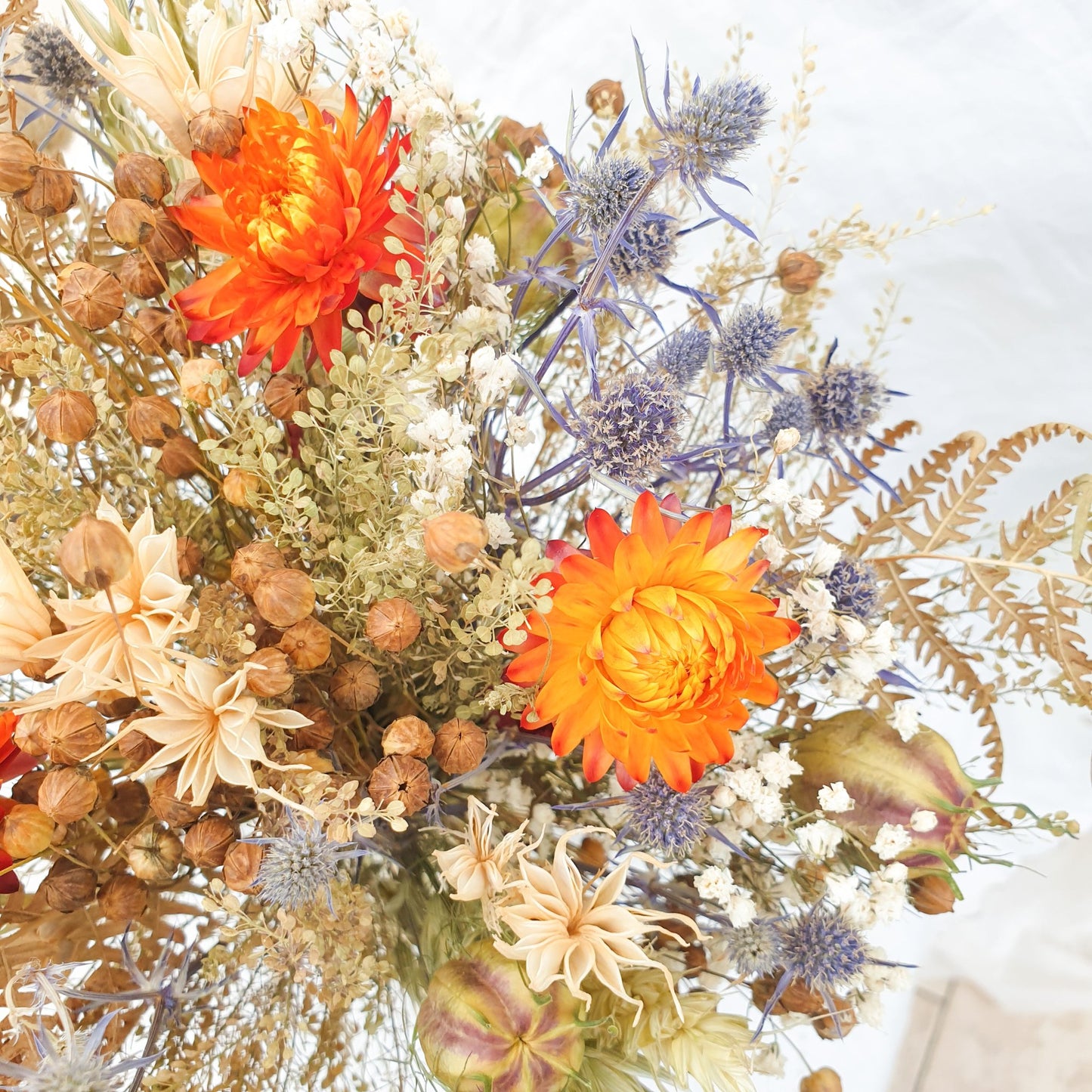 A close up of the smaller bouquet. You can see it contrains orange dried flowers contrasted with small blue thistles and fluffy bunny tail grasses along with green and brown filler stems and natural Welsh bracken foliage.