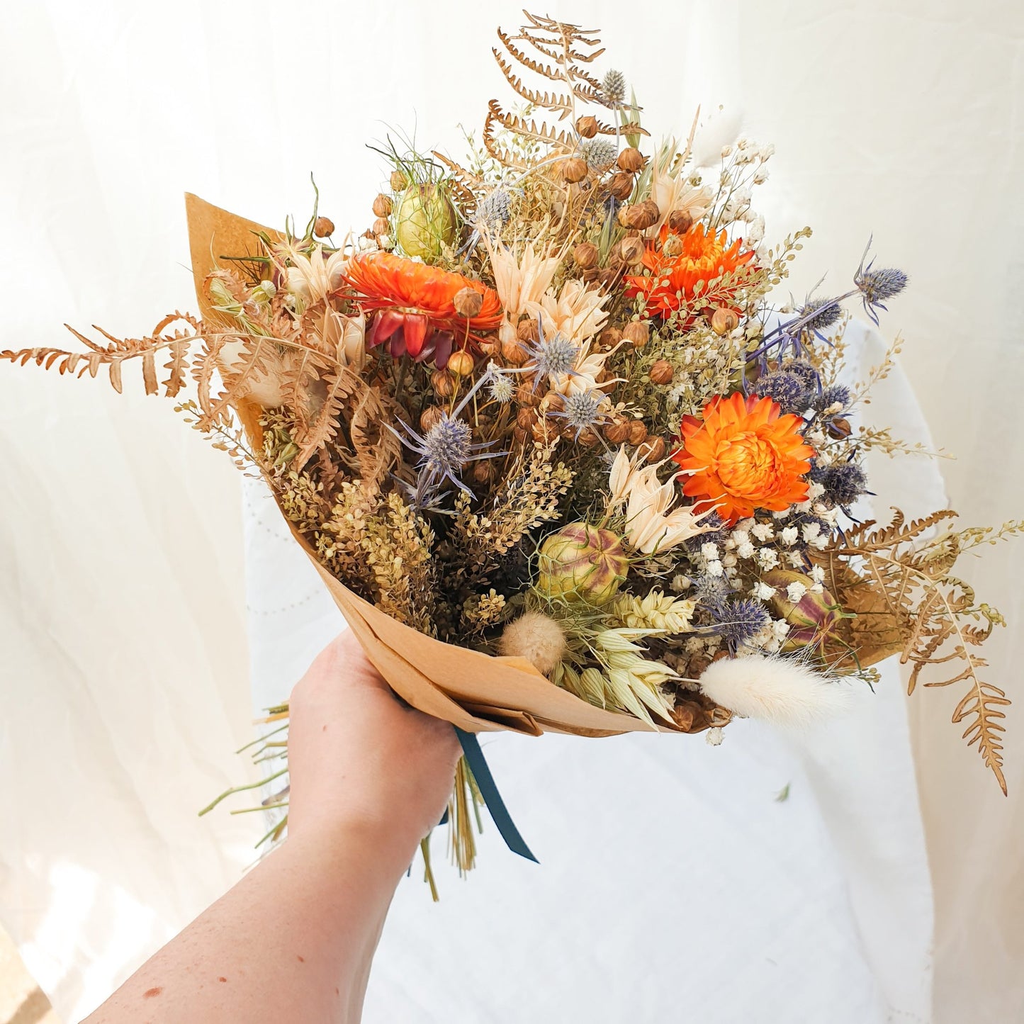A close up of the smaller bouquet wrapped stylishly in brown paper held in the hand. You can see orange dried flowers contrasted with small blue thistles and fluffy bunny tail grasses along with green and brown filler stems and natural Welsh bracken foliage.