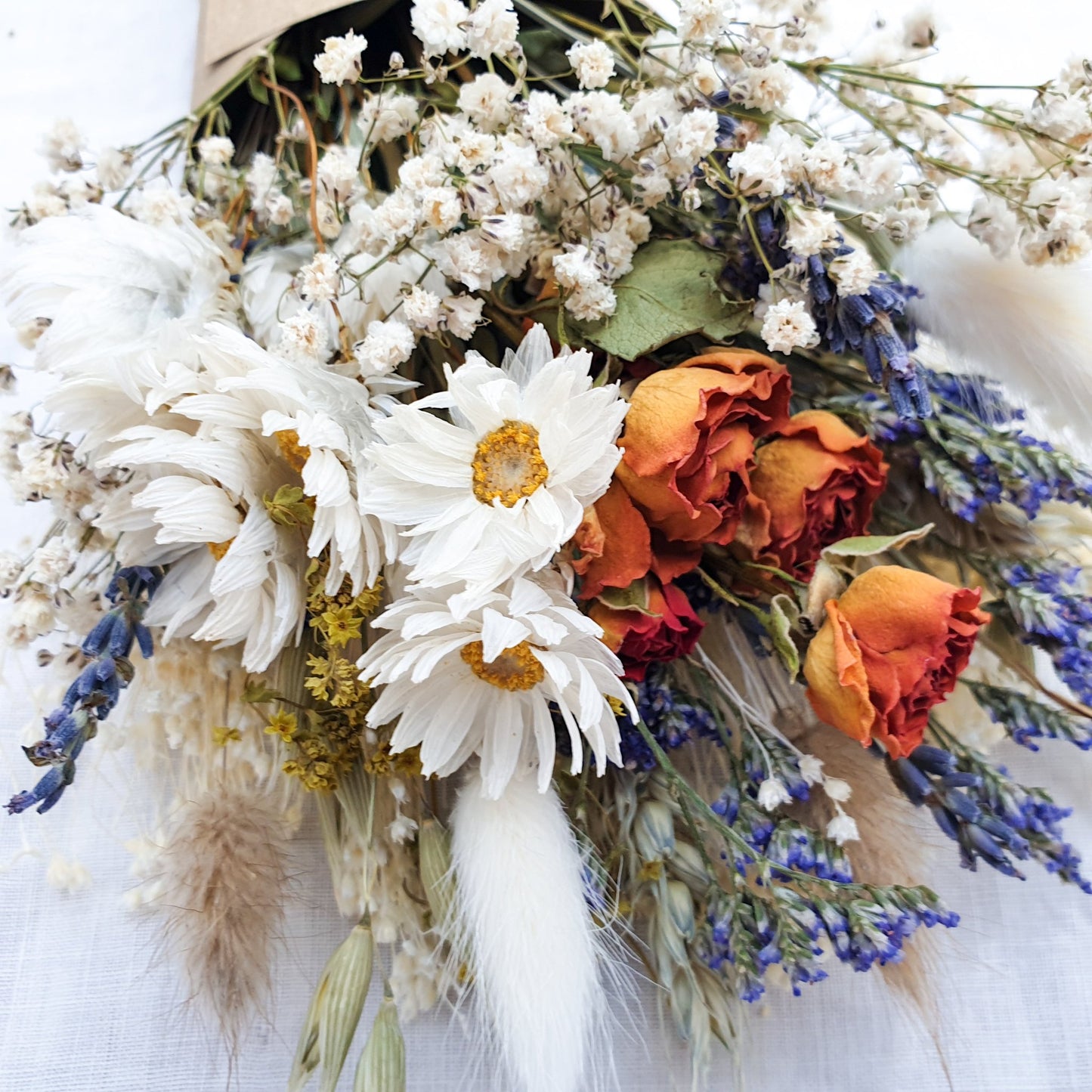 A close up of the dried flower bouquet. You can see in detail the orange spray roses, crisp white daisies with yellow centres, purple blue toned sea lavender and frothy white gypsophila among other grasses and fillers. You can see the pretty colouration of the orange roses, fading from a vibrant tomato red to orange to a golden apricot shade at the base.