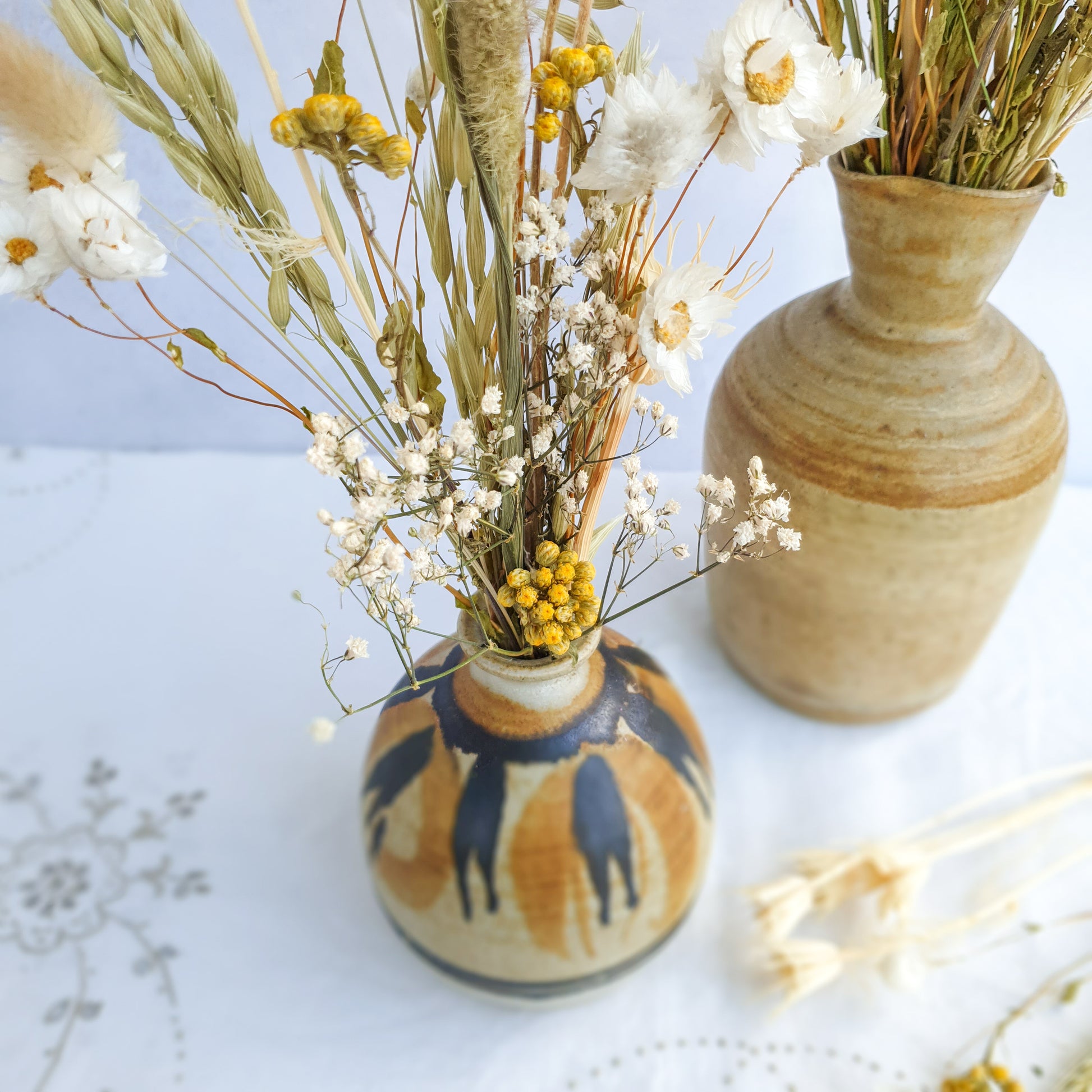 A brown ceramic vase filled with a vibrant bouquet of dried flowers. The bouquet features a mix of white daisies, yellow craspedia, and dried grasses, including lagurus fluffy bunny tail grass and wheat.