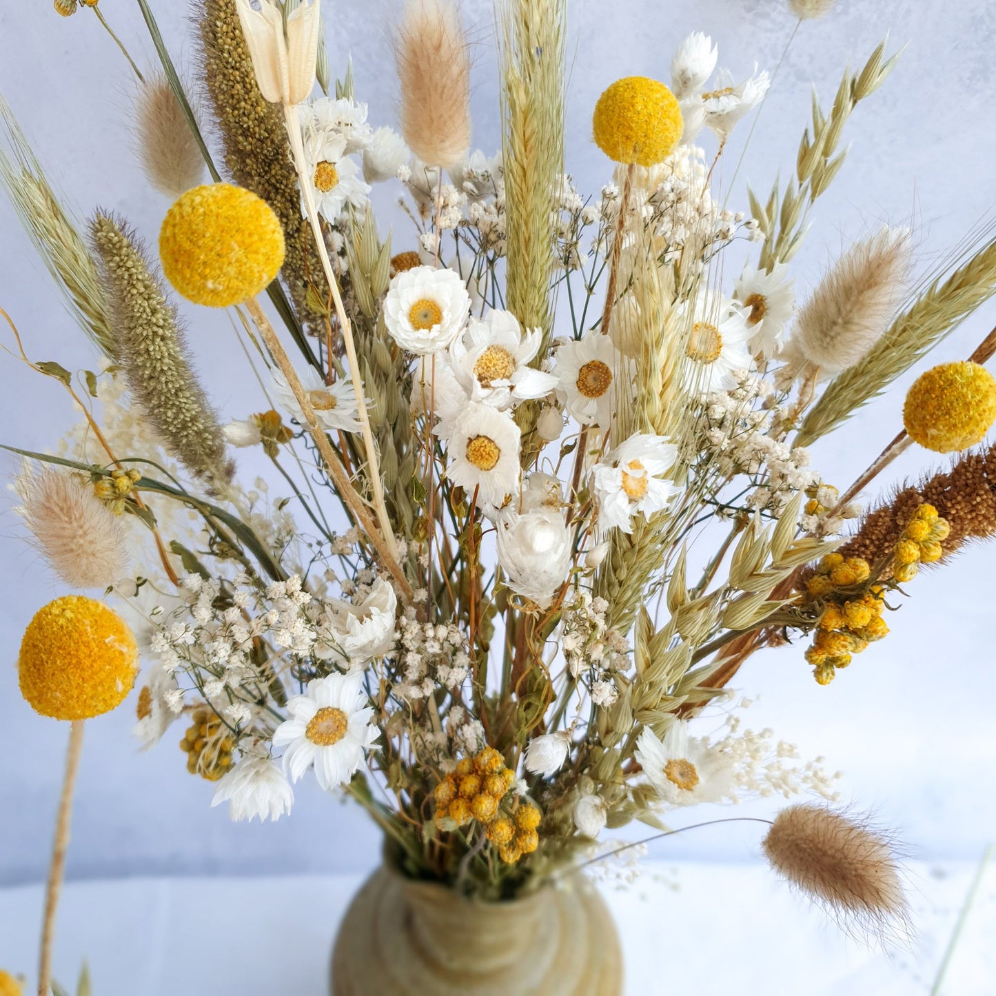 A brown ceramic vase filled with a vibrant bouquet of dried flowers. The bouquet features a mix of white daisies, yellow craspedia, and dried grasses, including lagurus fluffy bunny tail grass and wheat.