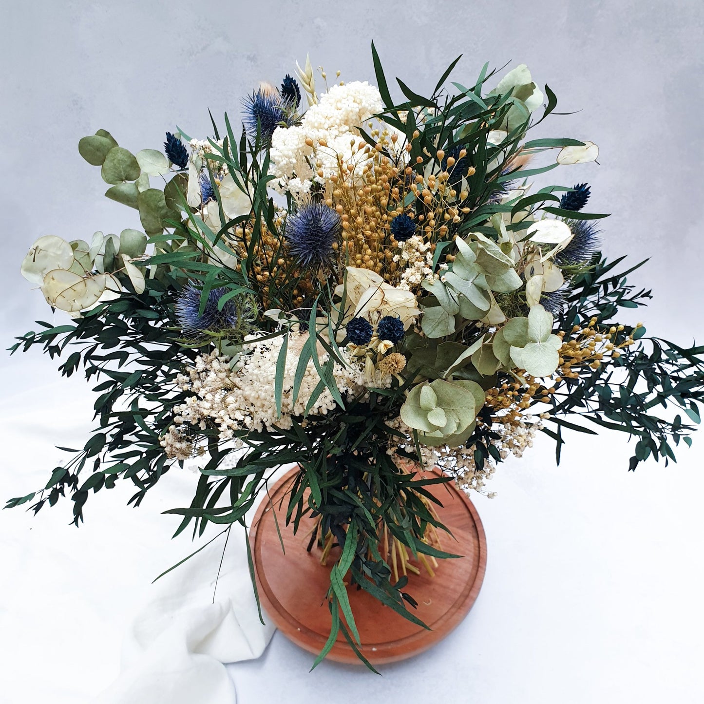 "Frosted Forest" Dried and Preserved Eucalyptus Bouquet - Lytton Rose Botanical. A beautifully arranged bouquet of dried and preserved foliages, showcasing a variety of textures and colors. Delicate white flowers, vibrant blue thistles, and lush green eucalyptus leaves create a stunning contrast. The bouquet is elegantly displayed on a rustic wooden board.