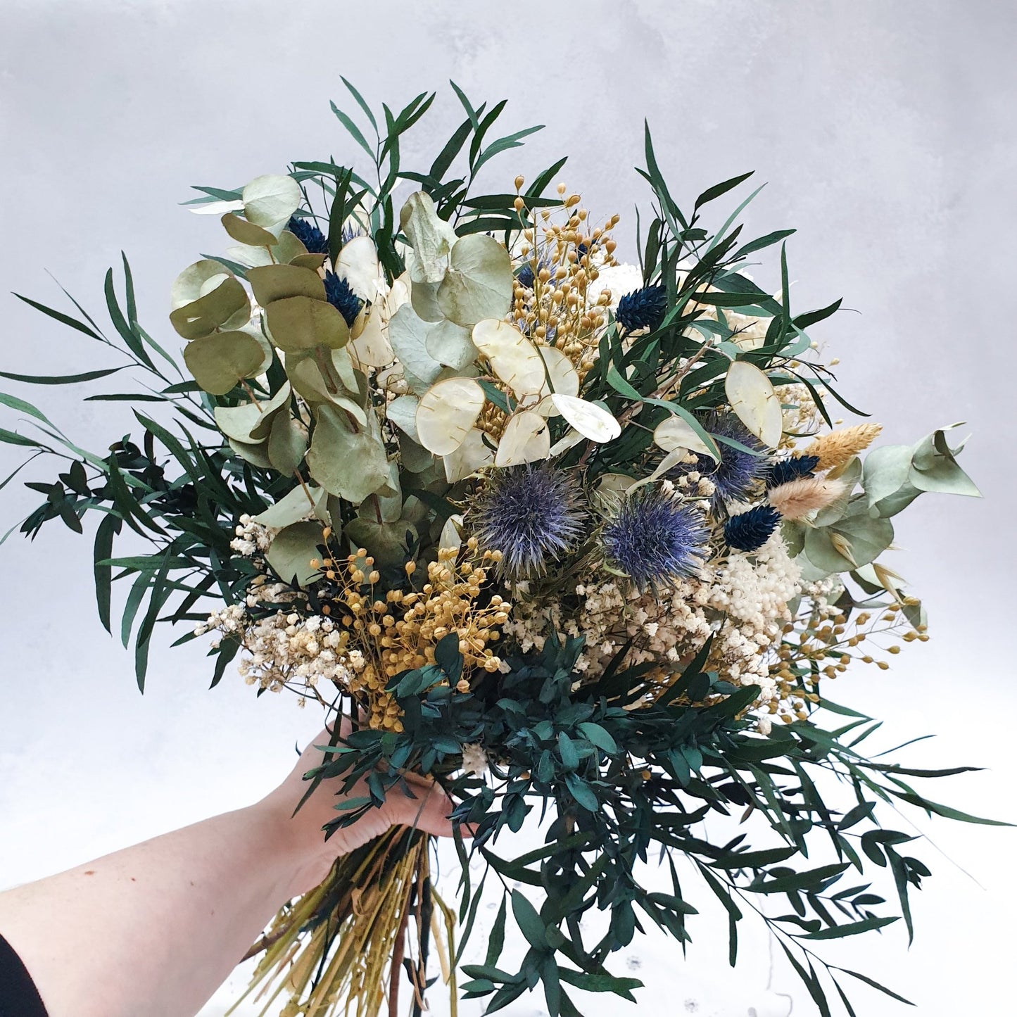 "Frosted Forest" Dried and Preserved Eucalyptus Bouquet - Lytton Rose Botanical. A beautifully arranged bouquet of dried and preserved foliage, showcased by a hand holding the arrangement. Delicate white flowers, vibrant blue thistles, and lush green eucalyptus leaves create a stunning contrast. The bouquet is tied with twine.