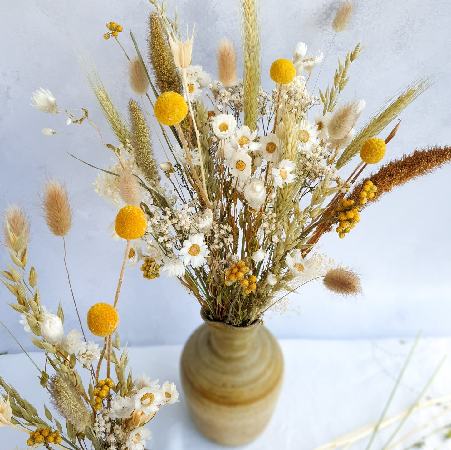 A brown ceramic vase filled with a vibrant bouquet of dried flowers. The bouquet features a mix of white daisies, yellow craspedia, and dried grasses, including lagurus fluffy bunny tail grass and wheat.