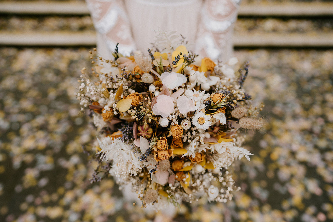 Close-up of a bride holding a rustic dried flower wedding bouquet with yellow roses, white daisies, lavender flowers, and wildflowers in sunny tones, captured outdoors with scattered floral confetti.