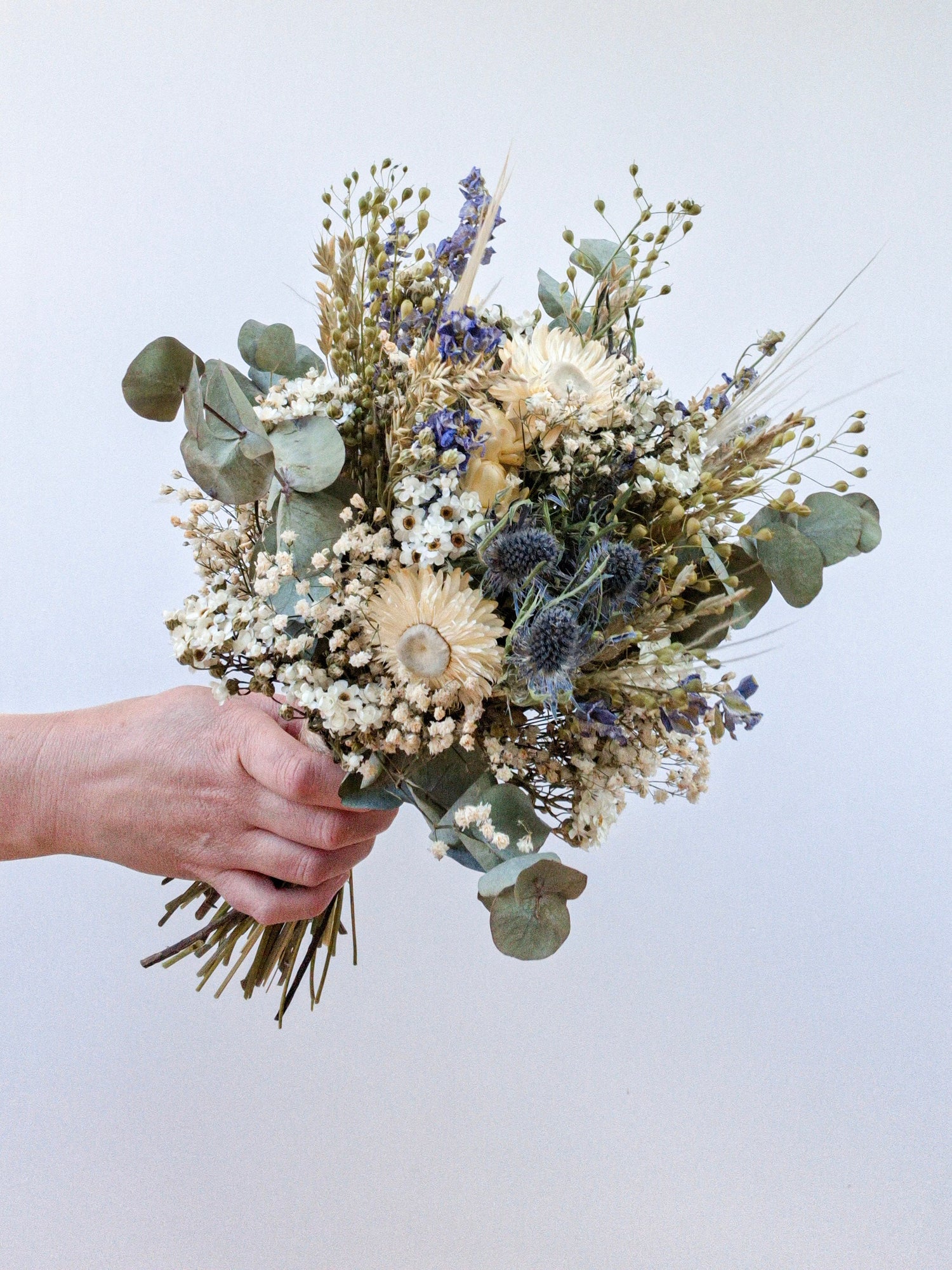 A hand holding a pretty blue and white dried flower bouquet against a plain white background, showing the natural stems of thistles, daisies, ivory strawflowers and eucalyptus, finished with baby's breath.