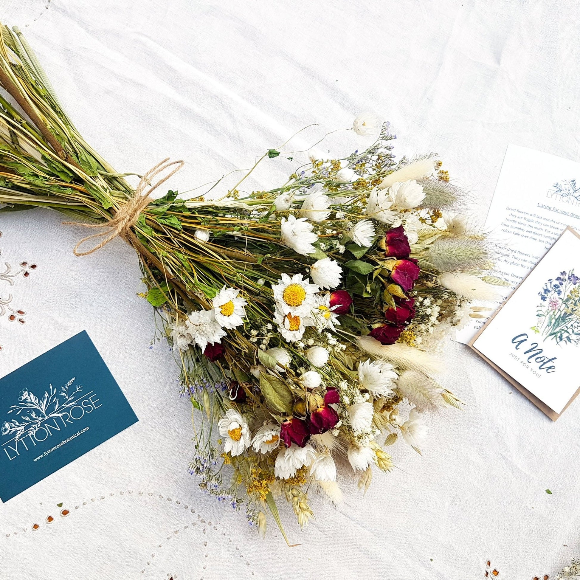 A dried flower bouquet is laid on a white linen background. It has small red roses, white daisies with yellow centres, blue toned purple sea lavender and pretty white gypsophila among other grasses and fillers. In the background you can see the optional gift card and instructions for caring for dried flowers. It is tied with a pretty twine binding.