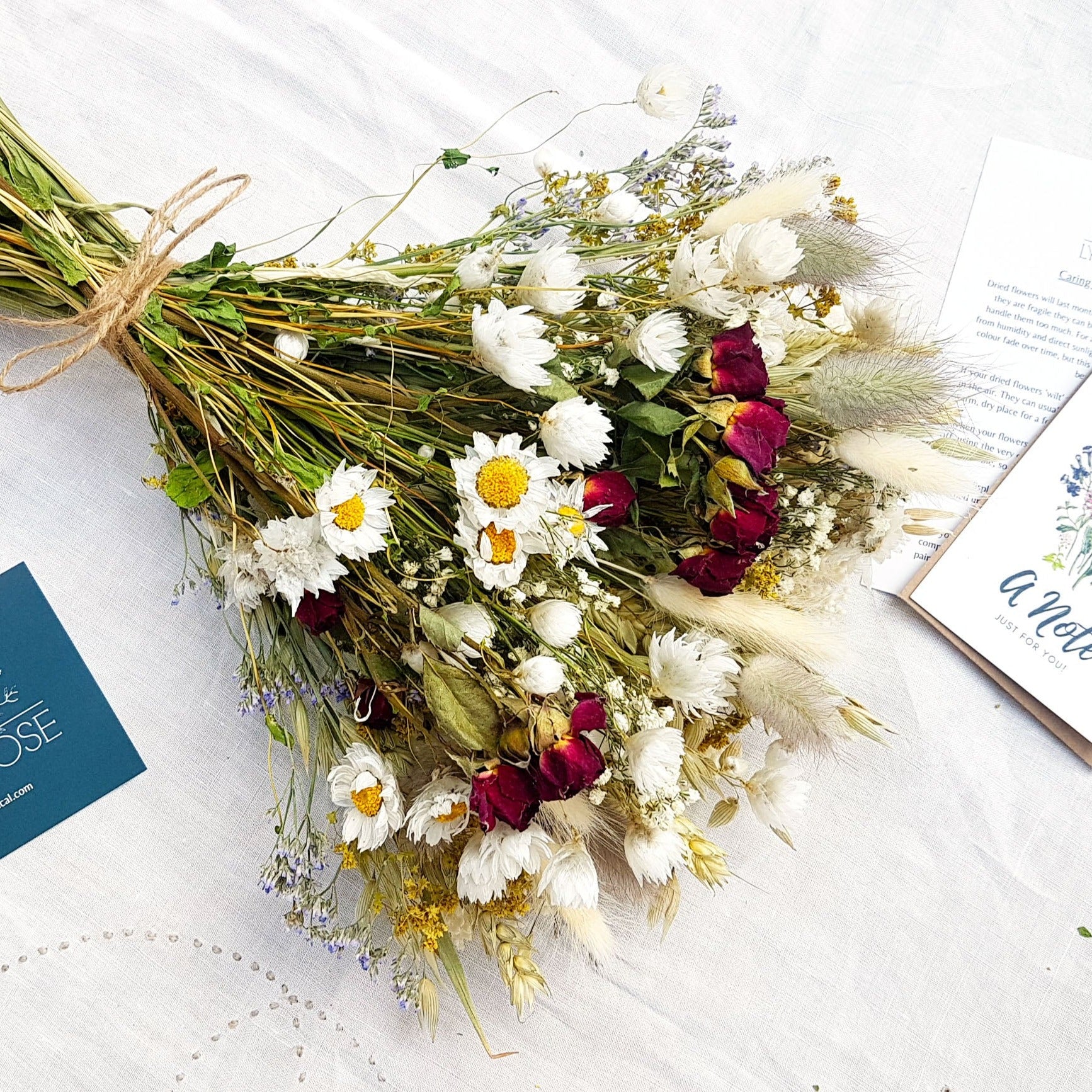 A close up image of the dried flower bouquet. It has small red roses, white daisies with yellow centres, blue toned purple sea lavender and pretty white gypsophila among other grasses and fillers. 