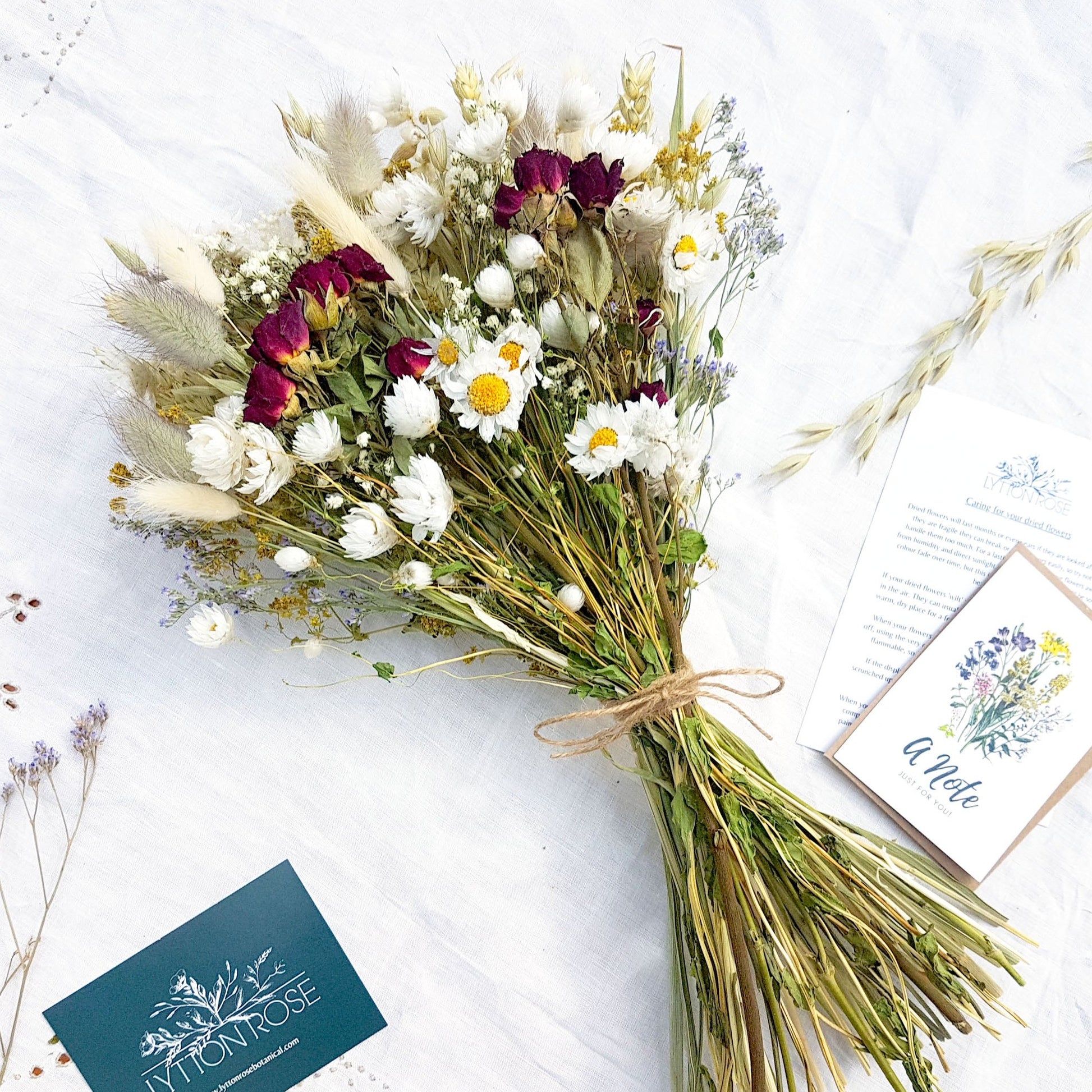 A dried flower bouquet is laid on a white linen background. It has small red roses, white daisies with yellow centres, blue toned purple sea lavender and pretty white gypsophila among other grasses and fillers. In the background you can see the optional gift card and instructions for caring for dried flowers. It is tied with a pretty twine binding.