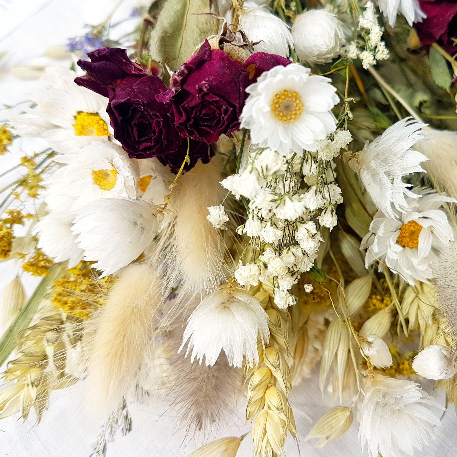 A close up of the dried flower bouquet. You can see in detail the red spray roses, crisp white daisies with yellow centres, purple blue toned sea lavender and frothy white gypsophila among other grasses and fillers. 