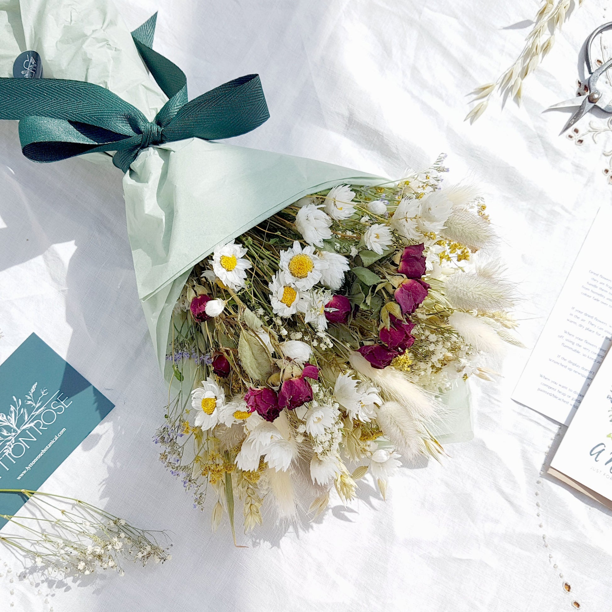 A dried flower bouquet wrapped in tissue is laid on a white linen cloth. It has small red roses, white daisies with yellow centres, lilac sea lavender and pretty white gypsophila among other grasses and fillers. In the background you can see the optional gift card and instructions for caring for dried flowers.