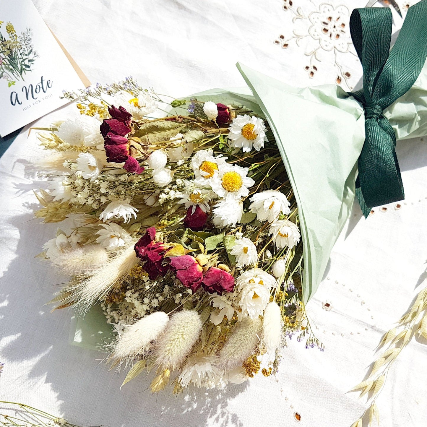 A dried flower bouquet wrapped in tissue is laid on a white linen cloth. It has small red roses, white daisies with yellow centres, lilac sea lavender and pretty white gypsophila among other grasses and fillers. In the background you can see the optional gift card and instructions for caring for dried flowers.