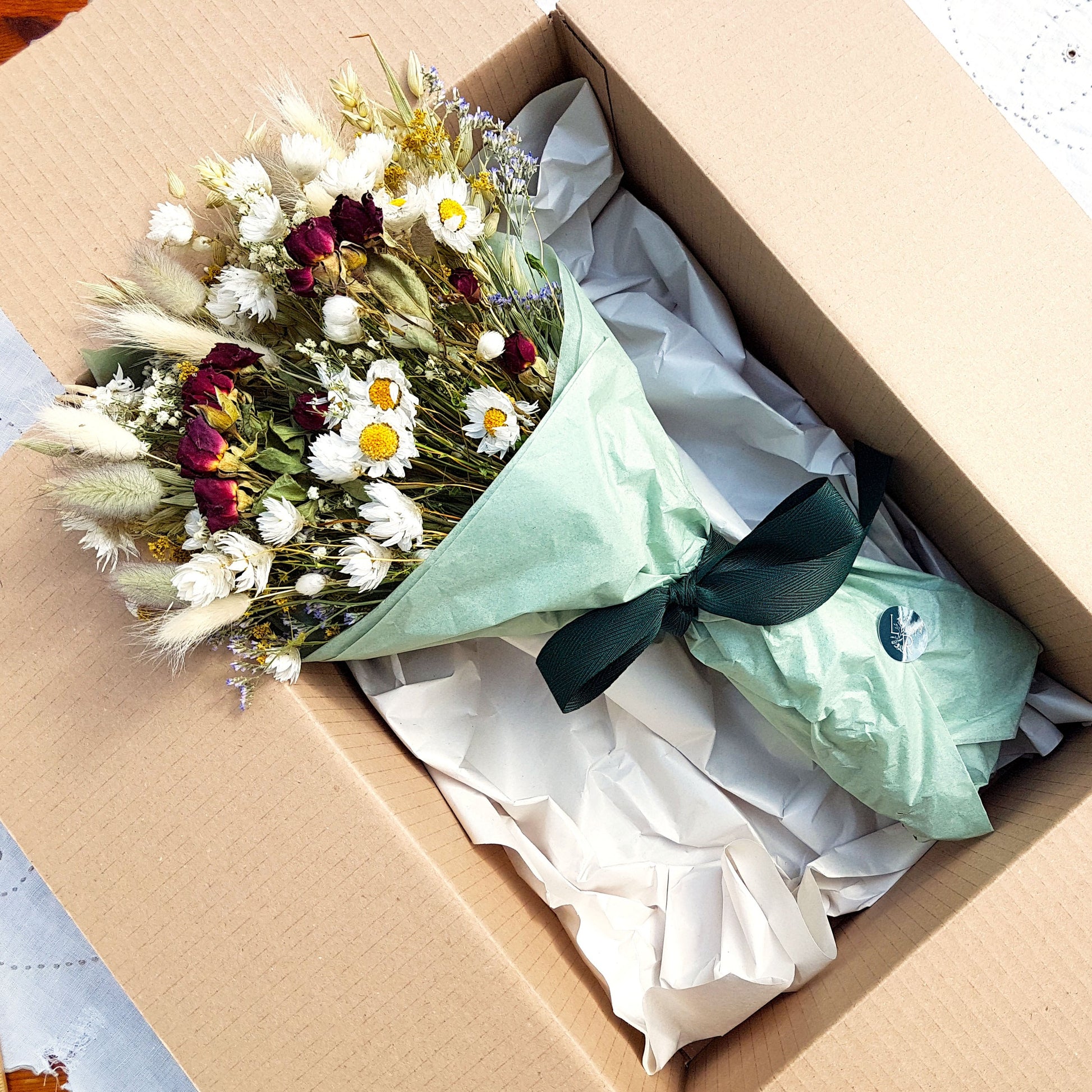 A close up of the dried flower bouquet wrapped sits in its packaging box with a pretty recycled tissue layer. It has small deep red roses, white daisies with yellow centres, lilac sea lavender and pretty white gypsophila among other grasses and fillers. In the background you can see the optional gift card and instructions for caring for dried flowers.