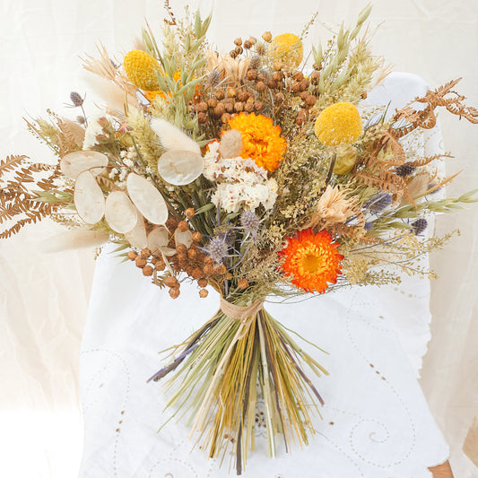 An Autumnal themed dried flower bouquet in a rounded handtie style. Orange and yellow dried flowers are accented with blue thistles, silvery honestly lunaria and fluffy bunny tail grasses. There are green and brown filler stems and natural Welsh bracken as foliage. The bouquet is informal, romantic and wild, as if it has been gathered together on a walk in the countryside. It is finished with a binding of rustic twine.