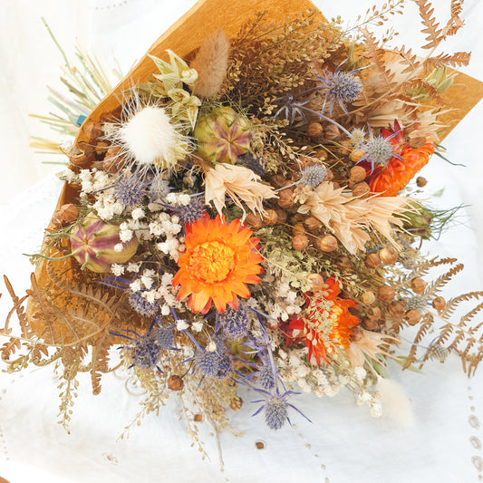 A close up of the smaller bouquet laid on one side showing all of the flowers it contains. You can see orange dried flowers contrasted with small blue thistles and fluffy bunny tail grasses along with green and brown filler stems and natural Welsh bracken foliage. It has a rounded yet informal and whimsical natural style and wrapped prettily in brown eco friendly wrapping.