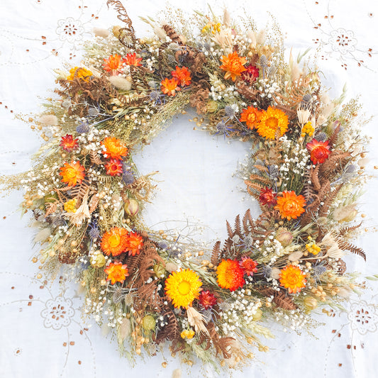 A wild, whimsical and natural looking dried flower wreath sits against a white background. The wreath has pops of vibrant orange and yellow strawflowers, delicate blue thistles and white fillers, such as frothy baby’s breath and structural nigella orientalis, with it’s star shaped seed heads. You can see bracken foliage and other green fillers such as oats and wheat.