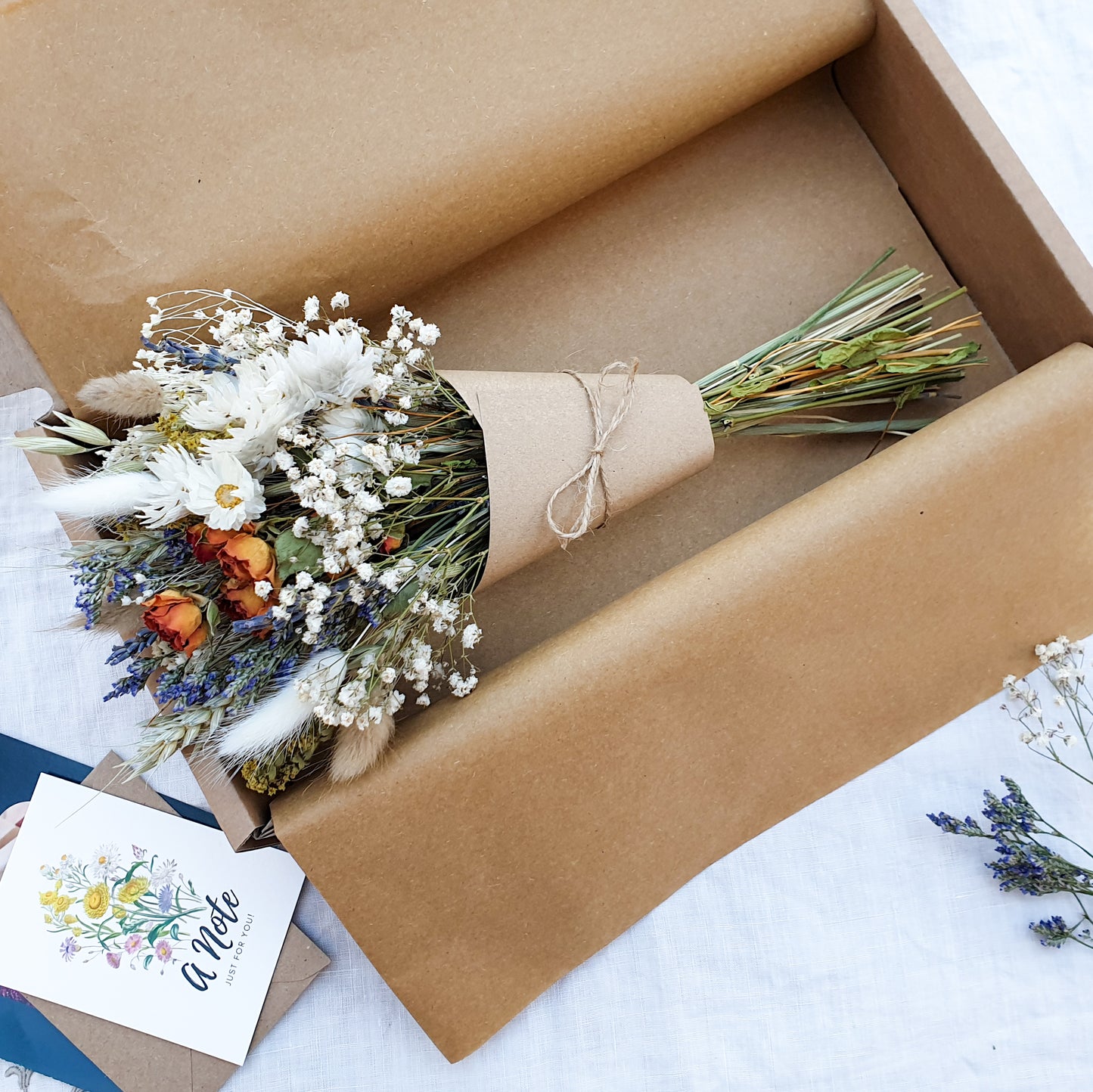 The dried flower bouquet wrapped in a kraft card cone sits in its packaging box with a pretty recycled tissue layer. It has small orange roses, white daisies with yellow centres, lilac sea lavender and pretty white gypsophila among other grasses and fillers. In the background you can see the optional gift card and instructions for caring for dried flowers.