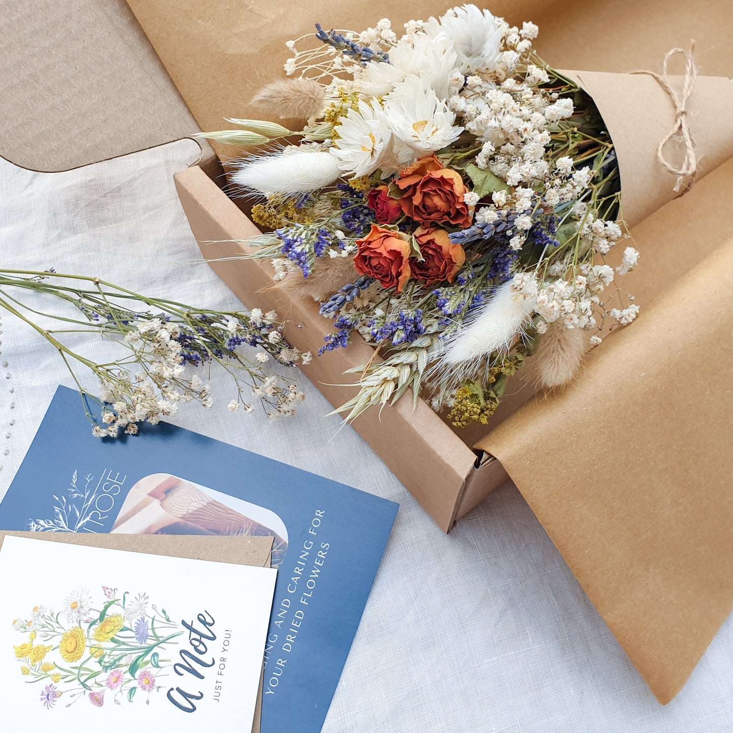 A close up of the dried flower bouquet wrapped sits in its packaging box with a pretty recycled tissue layer. It has small orange roses, white daisies with yellow centres, lilac sea lavender and pretty white gypsophila among other grasses and fillers. In the background you can see the optional gift card and instructions for caring for dried flowers. You can see the pretty colouration of the orange roses, fading from a vibrant tomato red to orange to a golden apricot shade at the base.
