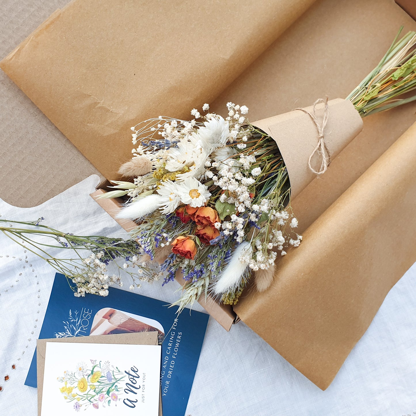 The dried flower bouquet wrapped in a kraft card cone sits in its packaging box with a pretty recycled tissue layer. It has small orange roses, white daisies with yellow centres, lilac sea lavender and pretty white gypsophila among other grasses and fillers. In the background you can see the optional gift card and instructions for caring for dried flowers.