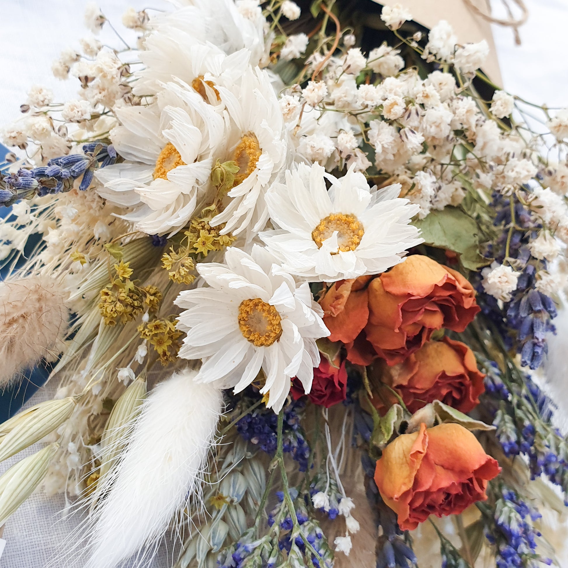 A close up of the dried flower bouquet. You can see in detail the orange spray roses, crisp white daisies with yellow centres, purple blue toned sea lavender and frothy white gypsophila among other grasses and fillers. You can see the pretty colouration of the orange roses, fading from a vibrant tomato red to orange to a golden apricot shade at the base.