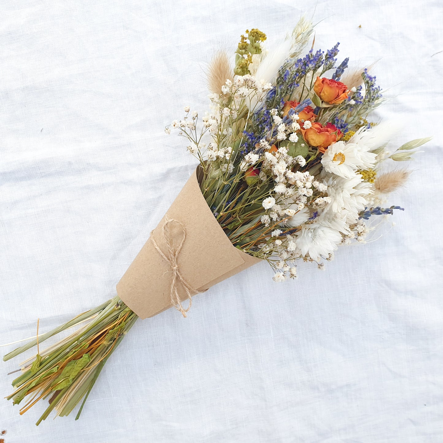 A dried flower bouquet wrapped in a kraft card cone is laid on a white linen cloth. It has small orange roses, white daisies with yellow centres, lilac sea lavender and pretty white gypsophila among other grasses and fillers. 