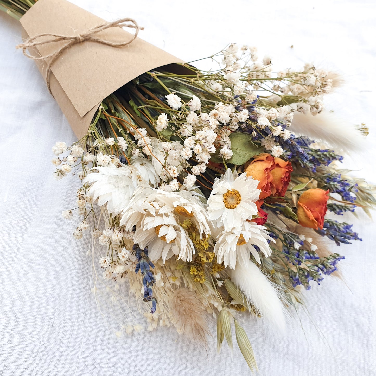 A dried flower bouquet wrapped in a kraft card cone is laid on a white linen cloth. It has small orange roses, white daisies with yellow centres, lilac sea lavender and pretty white gypsophila among other grasses and fillers. 