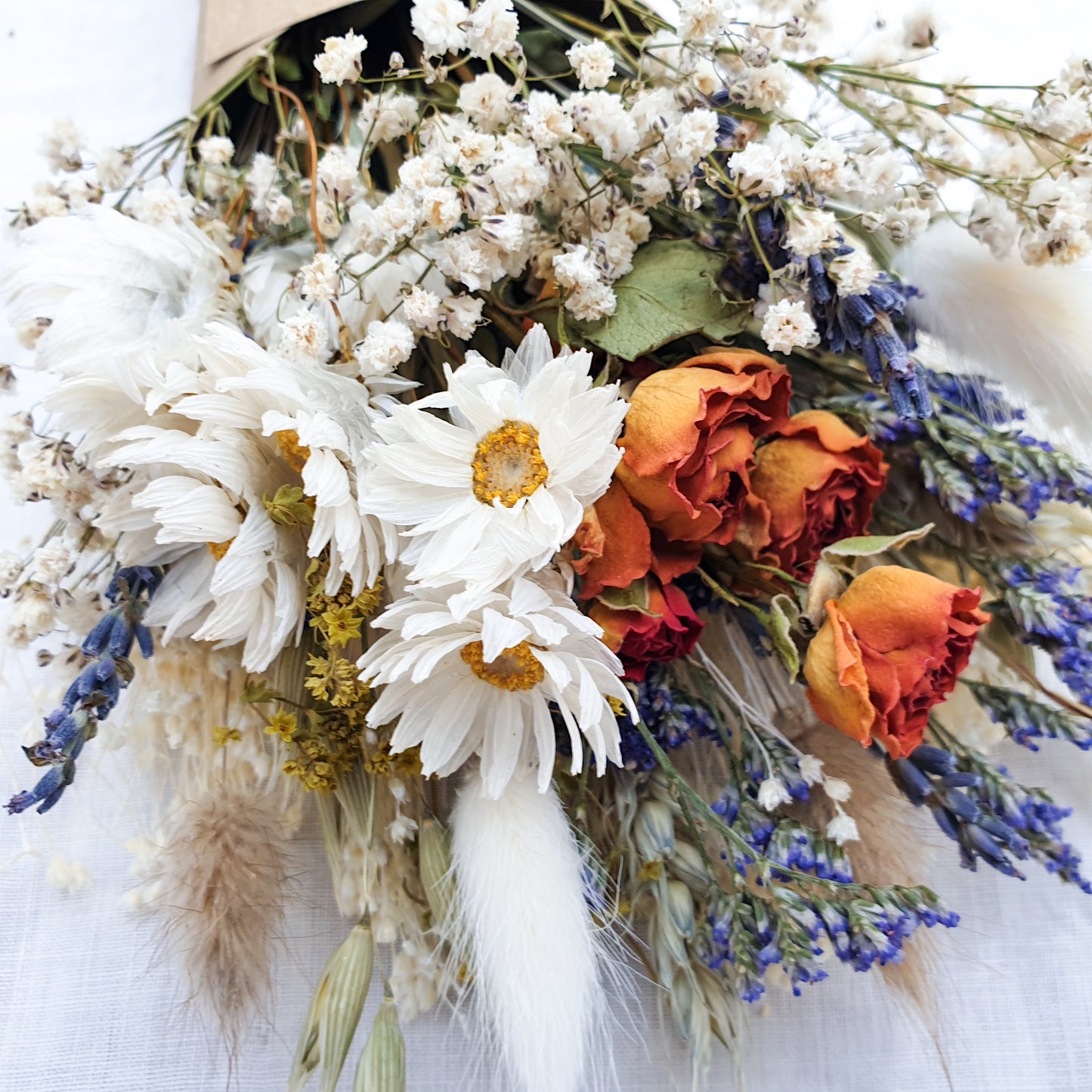 A close up of the dried flower bouquet. You can see in detail the orange spray roses, crisp white daisies with yellow centres, purple blue toned sea lavender and frothy white gypsophila among other grasses and fillers. You can see the pretty colouration of the orange roses, fading from a vibrant tomato red to orange to a golden apricot shade at the base. 