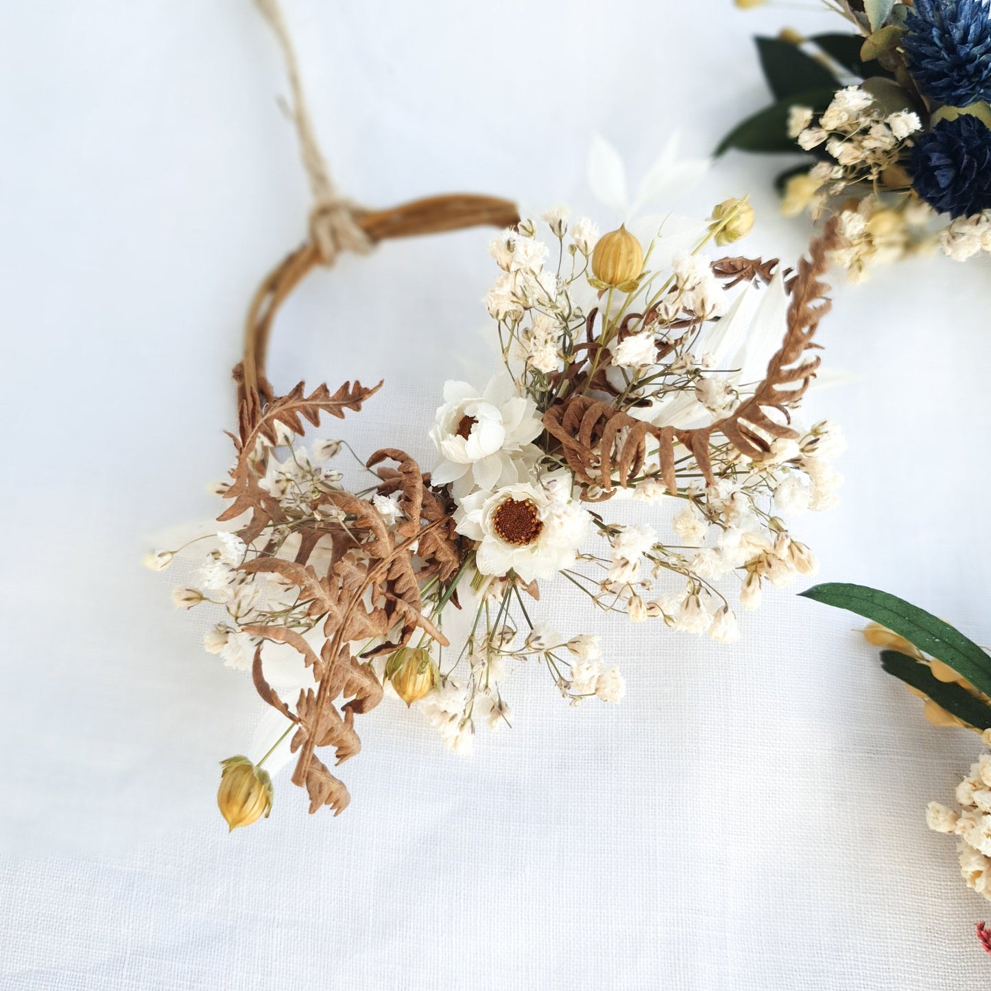 Mini Wreaths by Lytton Rose Botanical. A delicate, circular tiny wreath crafted from dried flowers and twigs. The wreath features a cluster of white dried flowers, including daisies and gypsophila, interspersed with dried brown ferns. The wreath is secured with a thin, natural-colored twine.