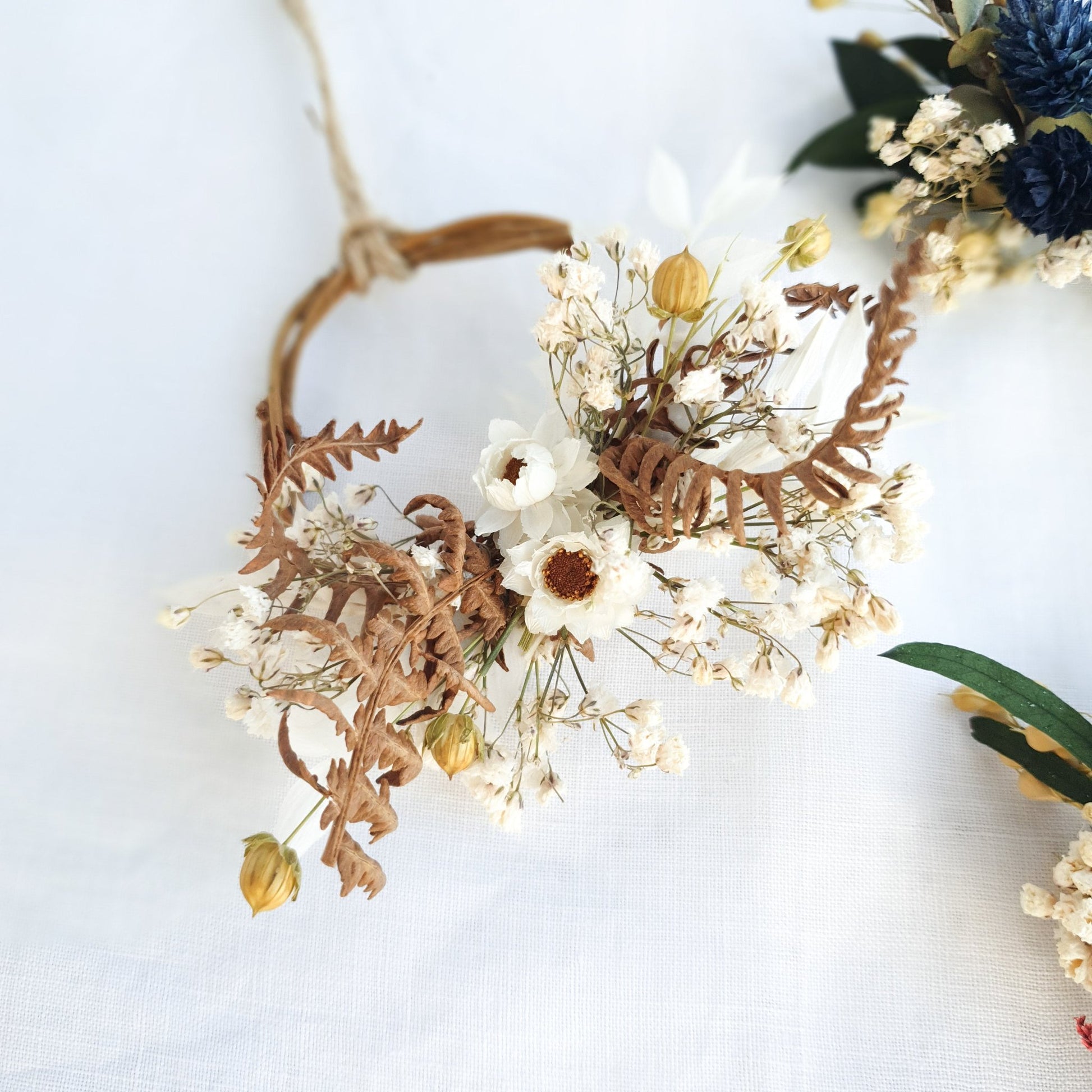 Mini Wreaths by Lytton Rose Botanical. A delicate, circular tiny wreath crafted from dried flowers and twigs. The wreath features a cluster of white dried flowers, including daisies and gypsophila, interspersed with dried brown ferns. The wreath is secured with a thin, natural-colored twine.