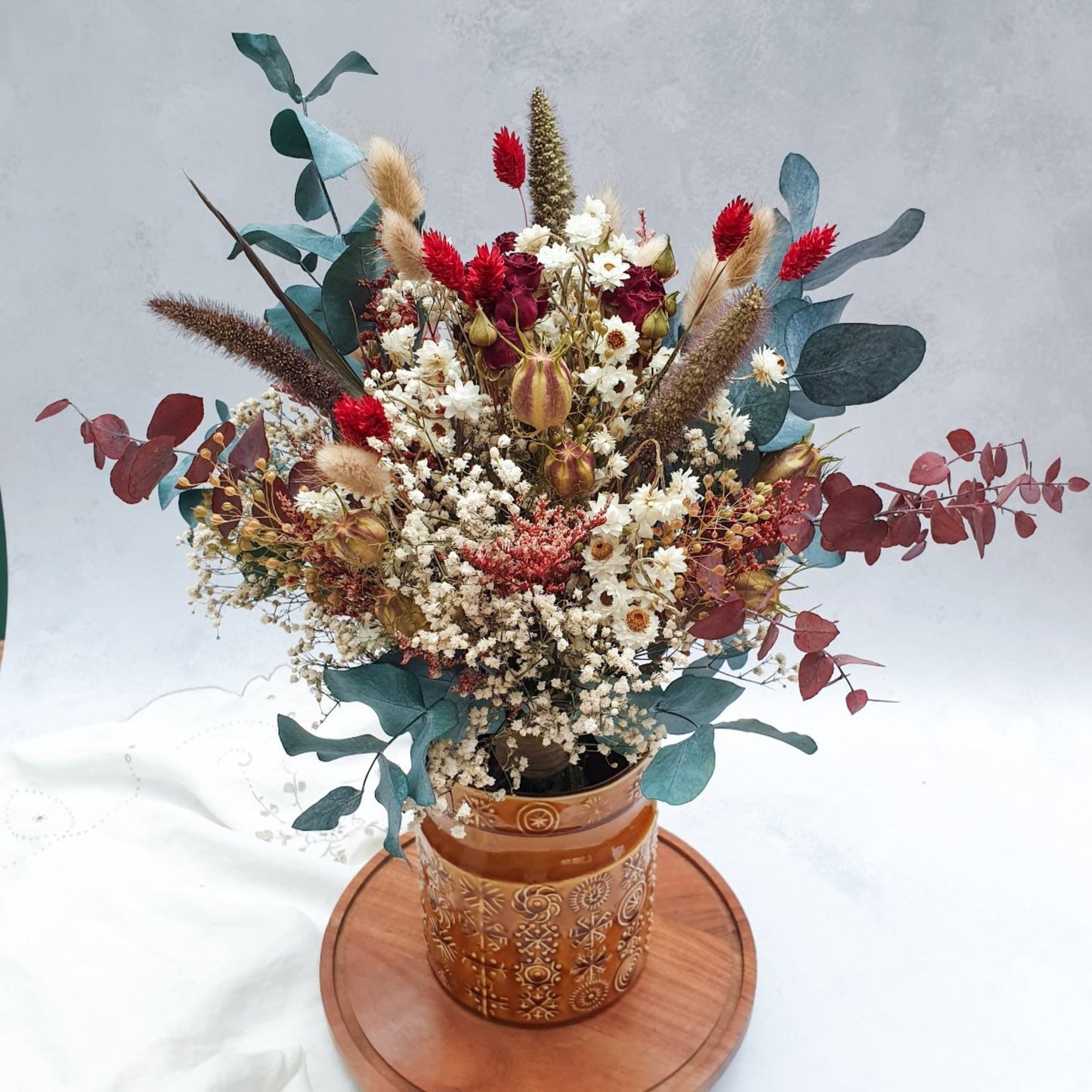 "Winter Woodlands" Preserved Eucalyptus & Dried Roses Bouquet - Lytton Rose Botanical. A brown ceramic vase (not included) filled with a vibrant bouquet of dried flowers. The bouquet features a mix of white gypsophila, red roses, burgundy and green eucalyptus leaves. The vase is displayed on a wooden tray.