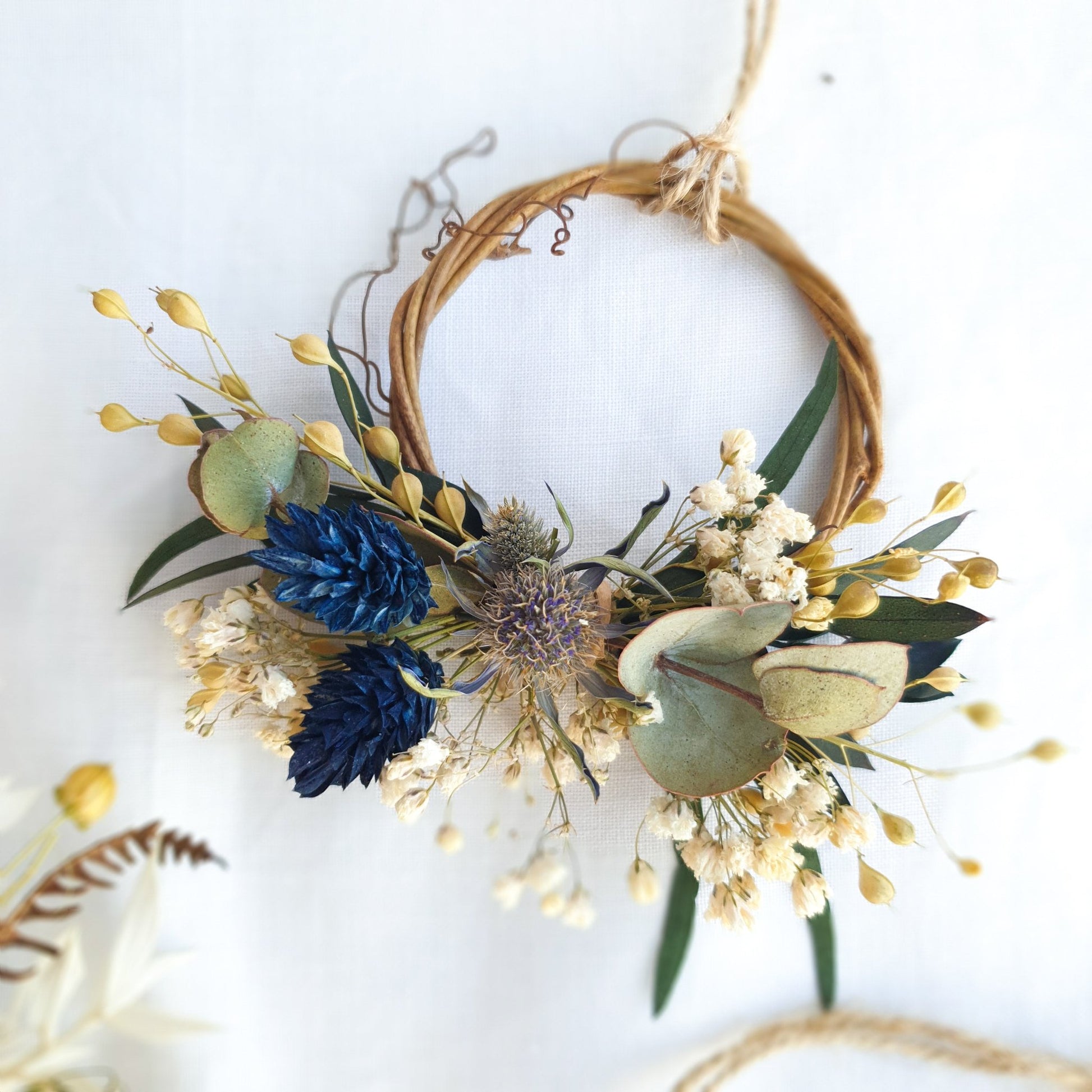Mini Wreath by Lytton Rose Botanical. A delicate, circular tiny wreath crafted from dried flowers and twigs. The wreath features a cluster of cream-colored dried flowers, including gypsophila and statice, interspersed with vibrant blue thistle-like flowers and lush green eucalyptus leaves. The wreath is secured with a thin, natural-colored twine.
