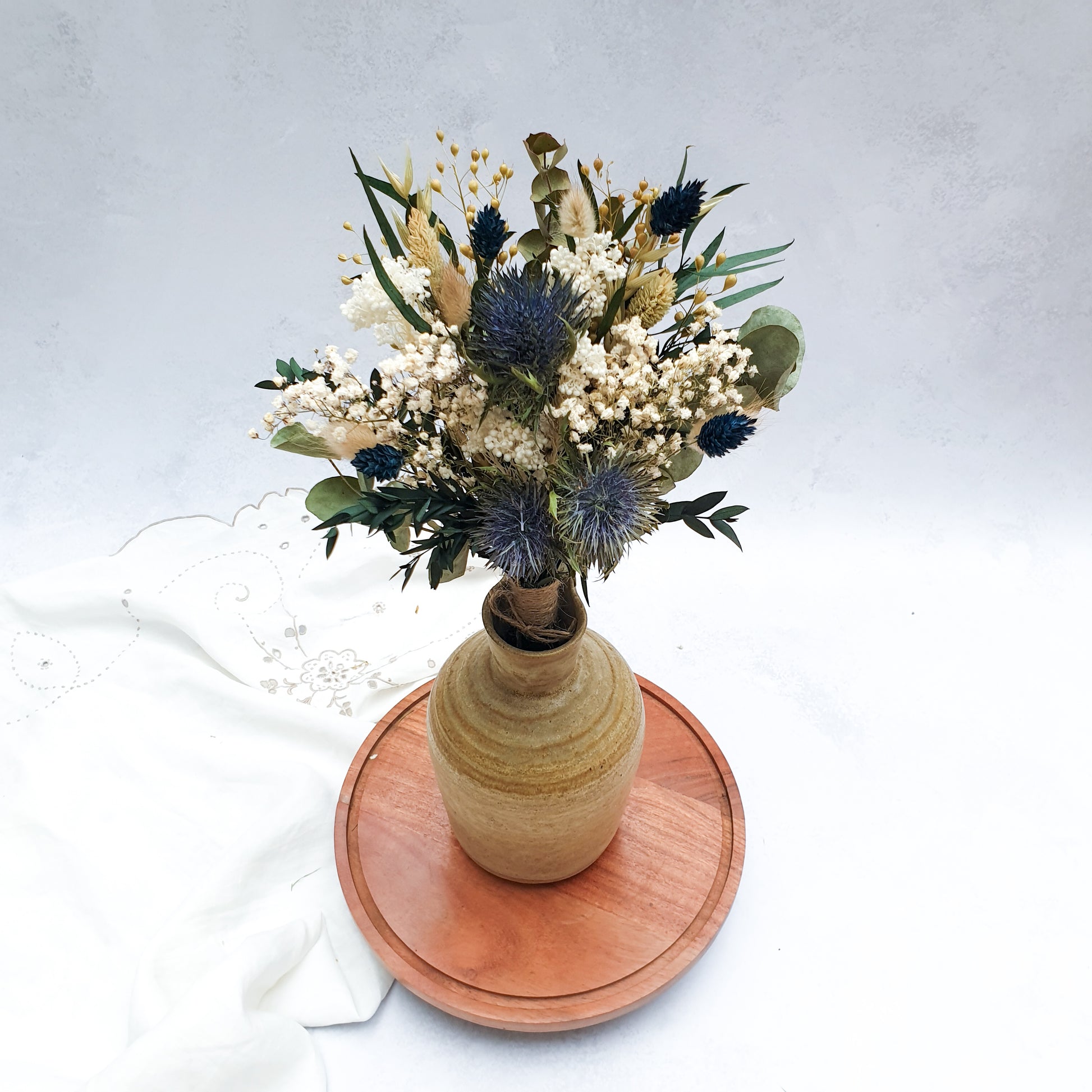 "Frosted Forest" Dried and Preserved Small Eucalyptus Bouquet - Lytton Rose Botanical. A brown ceramic vase filled with a vibrant bouquet of dried flowers. The bouquet features a mix of white gypsophila, blue thistles, and green eucalyptus leaves. The vase is displayed on a wooden tray.