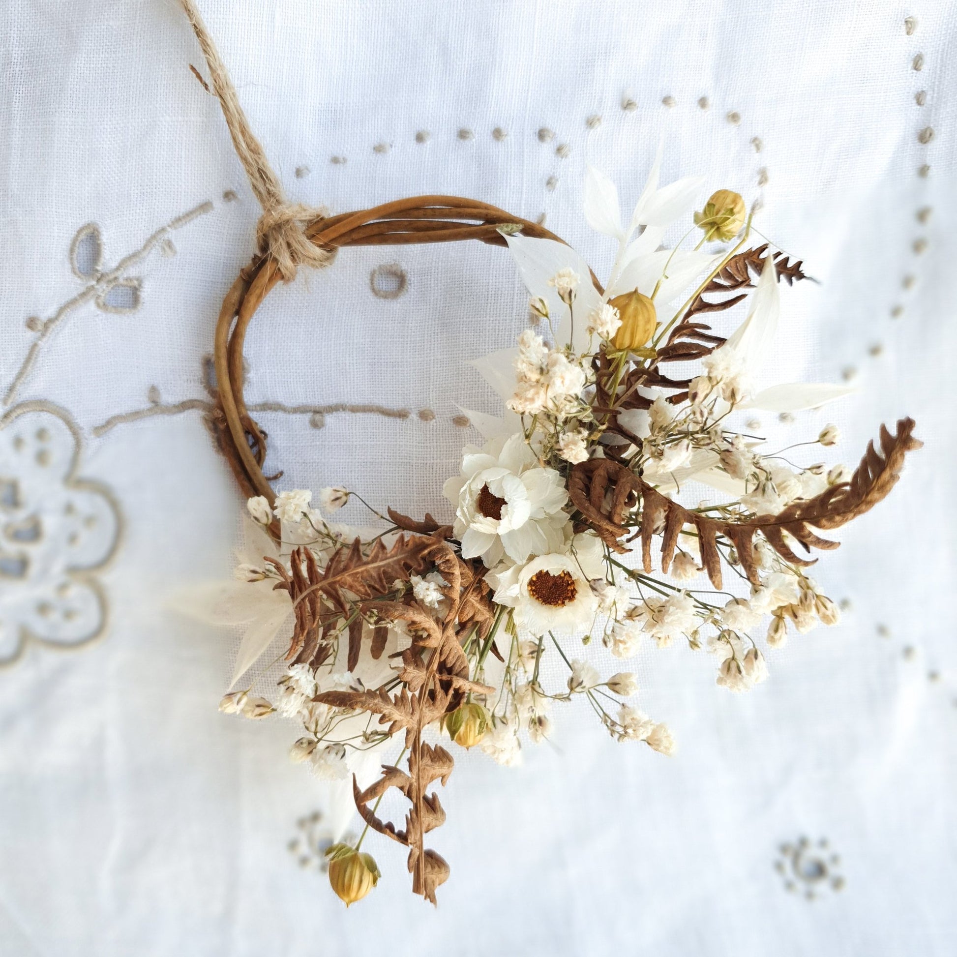 Mini Wreaths by Lytton Rose Botanical. A delicate, circular tiny wreath crafted from dried flowers and twigs. The wreath features a cluster of white dried flowers, including daisies and gypsophila, interspersed with dried brown ferns. The wreath is secured with a thin, natural-colored twine.