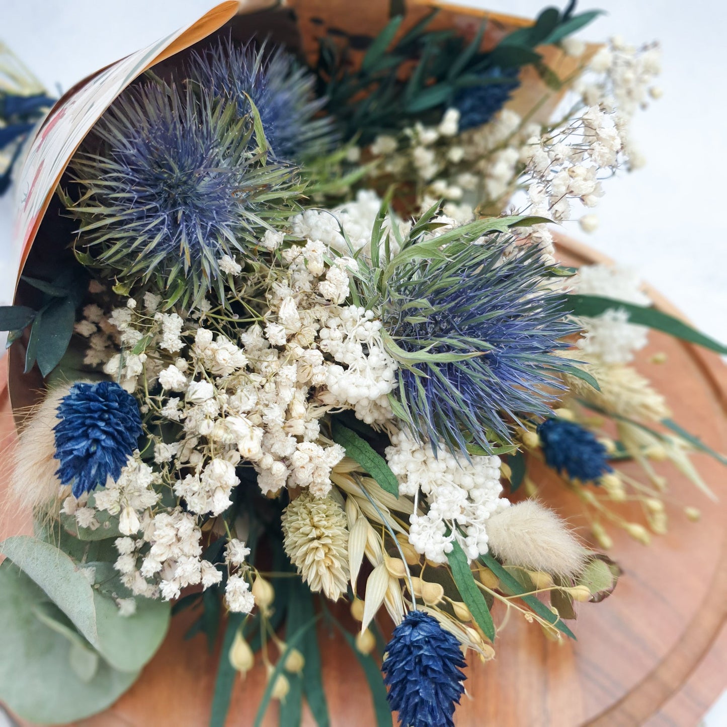 "Frosted Forest" Dried and Preserved Small Eucalyptus Bouquet - Lytton Rose Botanical. A beautifully arranged bouquet of dried flowers, wrapped in brown paper. Delicate white flowers, vibrant blue thistles, and lush green eucalyptus leaves create a stunning contrast.