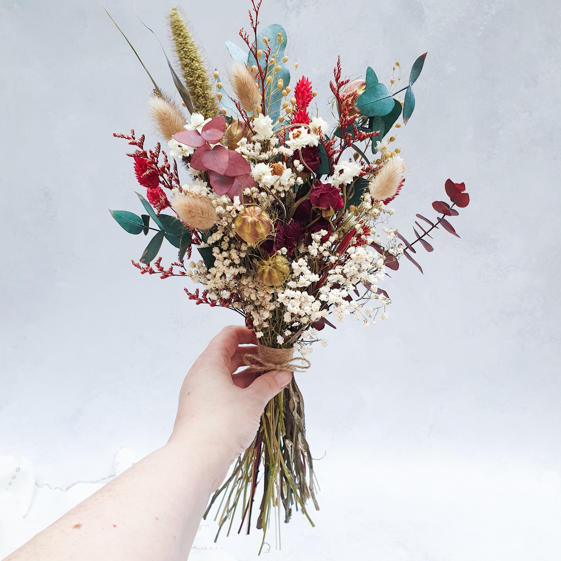 "Winter Woodlands" Preserved Eucalyptus & Dried Roses Small Bouquet - Lytton Rose Botanical. A beautifully arranged bouquet of dried flowers, showcasing a mix of white gypsophila, red roses, red and green eucalyptus leaves. The bouquet is elegantly tied with twine and is being held in a hand.