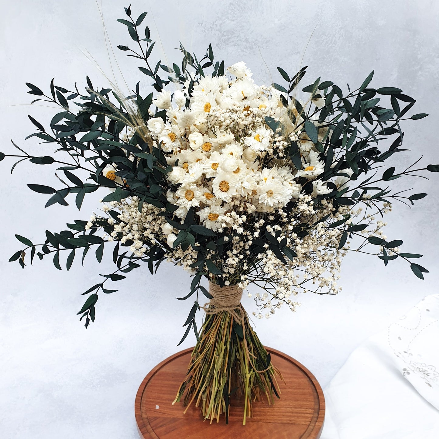 "Snowdrop Glade" Minimalist Large Handtie Bouquet - Lytton Rose Botanical. "Snowdrop Glade" Minimalist Large Handtie Bouquet - Lytton Rose Botanical. A beautifully arranged bouquet of dried flowers. Delicate white daisies are the centerpiece, surrounded by lush green eucalyptus leaves and wispy baby's breath. The bouquet is elegantly tied with twine and displayed on a rustic wooden board.
