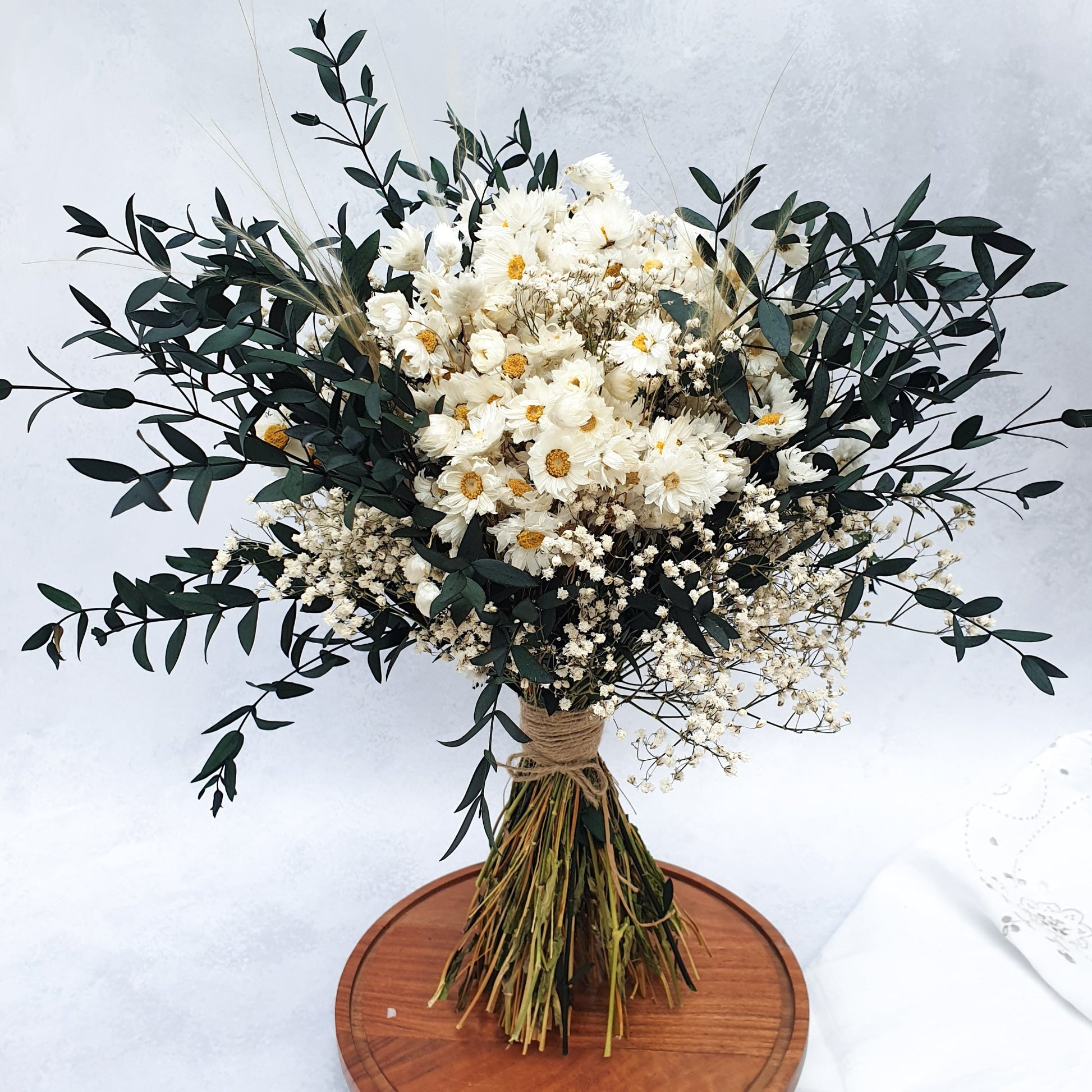 "Snowdrop Glade" Minimalist Large Handtie Bouquet - Lytton Rose Botanical. "Snowdrop Glade" Minimalist Large Handtie Bouquet - Lytton Rose Botanical. A beautifully arranged bouquet of dried flowers. Delicate white daisies are the centerpiece, surrounded by lush green eucalyptus leaves and wispy baby's breath. The bouquet is elegantly tied with twine and displayed on a rustic wooden board.