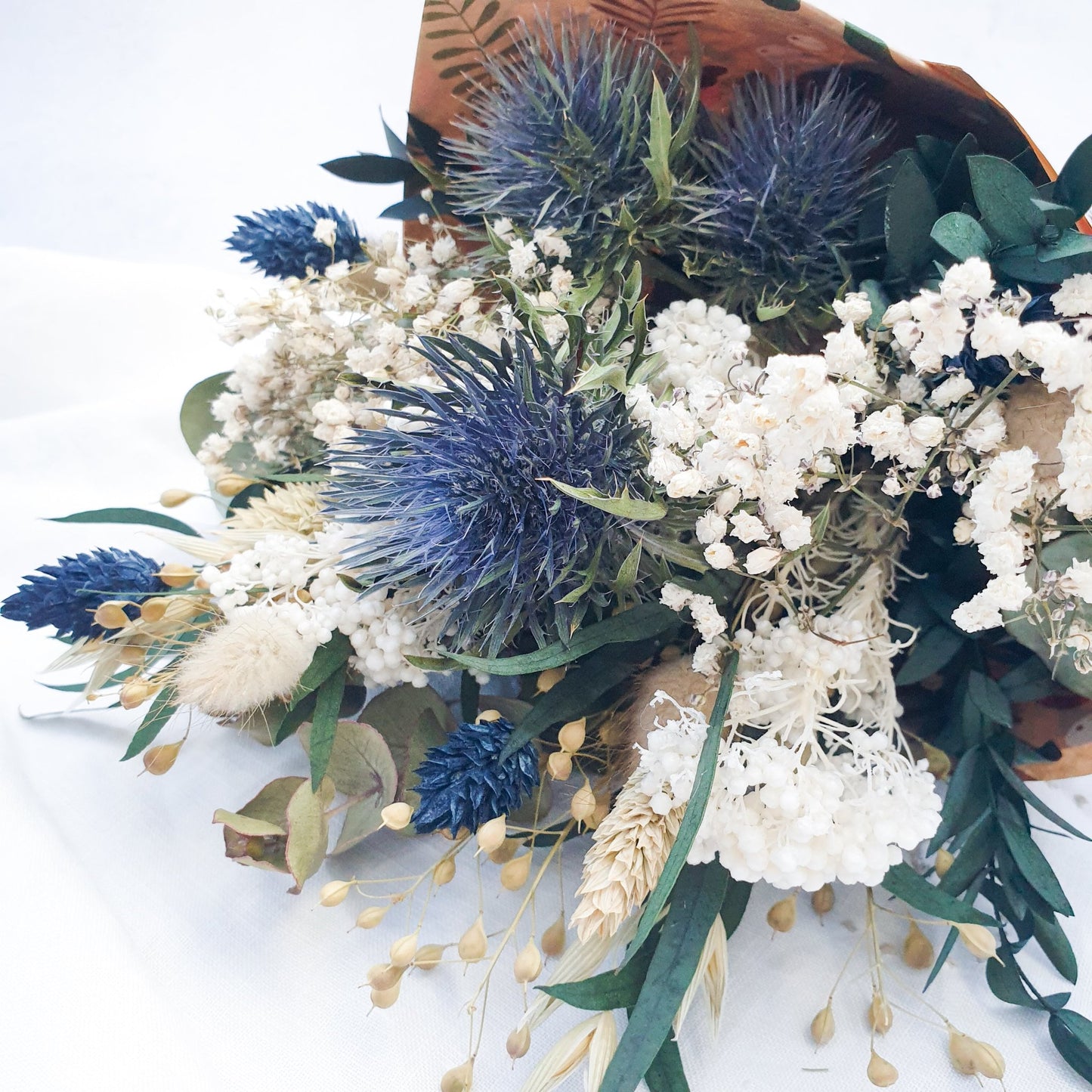 "Frosted Forest" Dried and Preserved Small Eucalyptus Bouquet - Lytton Rose Botanical. A beautifully arranged bouquet of dried flowers, wrapped in brown paper. Delicate white flowers, vibrant blue thistles, and lush green eucalyptus leaves create a stunning contrast.