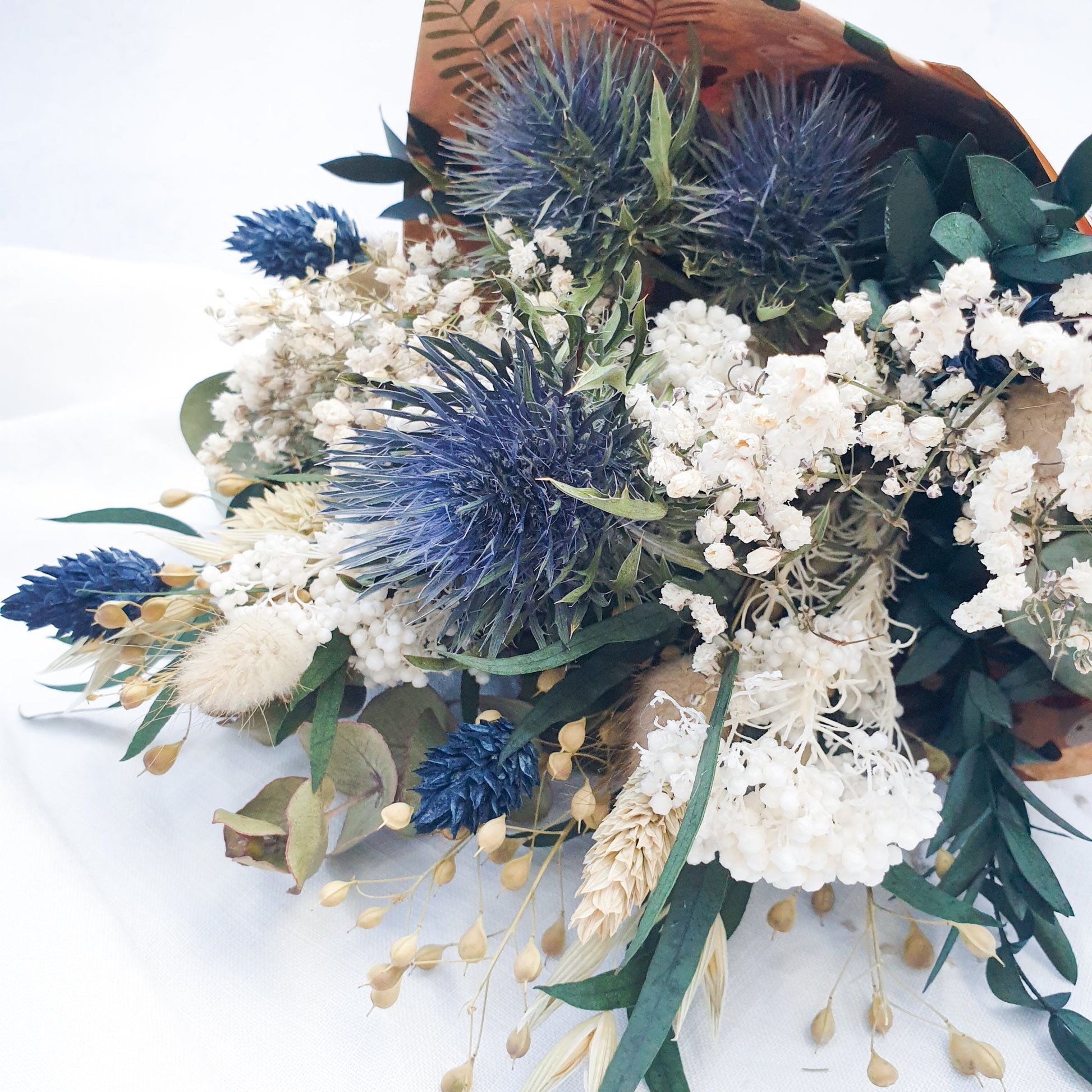 "Frosted Forest" Dried and Preserved Small Eucalyptus Bouquet - Lytton Rose Botanical. A beautifully arranged bouquet of dried flowers, wrapped in brown paper. Delicate white flowers, vibrant blue thistles, and lush green eucalyptus leaves create a stunning contrast.