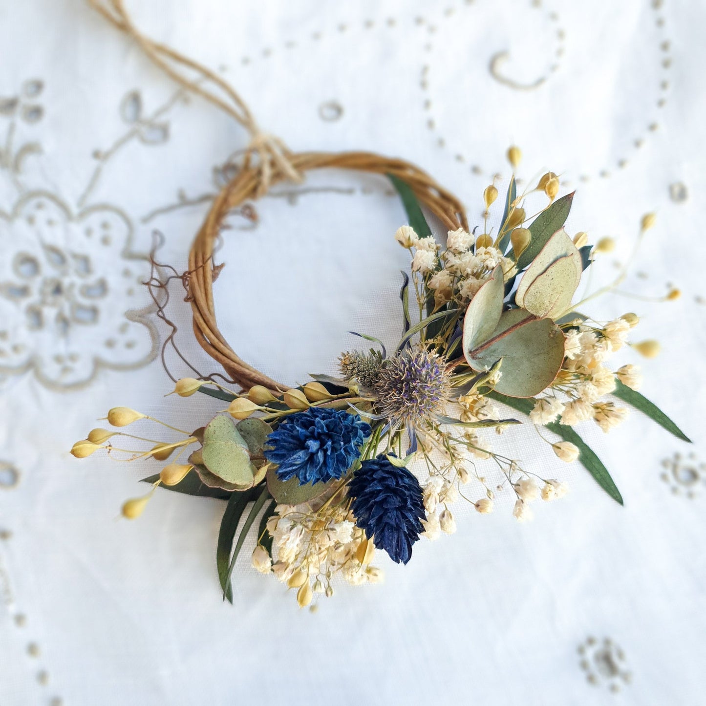 Mini Wreath by Lytton Rose Botanical. A delicate, circular tiny wreath crafted from dried flowers and twigs. The wreath features a cluster of cream-colored dried flowers, including gypsophila and statice, interspersed with vibrant blue thistle-like flowers and lush green eucalyptus leaves. The wreath is secured with a thin, natural-colored twine.