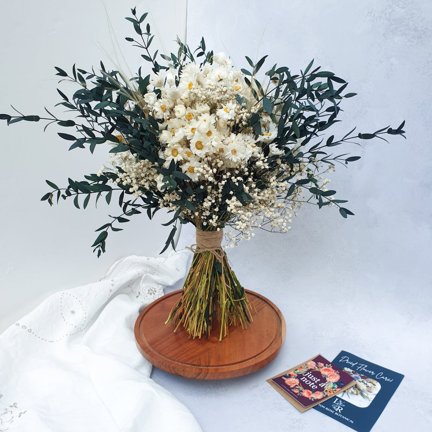 "Snowdrop Glade" Minimalist Large Handtie Bouquet - Lytton Rose Botanical. A beautifully arranged bouquet of dried flowers. Delicate white daisies are the centerpiece, surrounded by lush green eucalyptus leaves and wispy baby's breath. The bouquet is elegantly tied with twine and displayed on a rustic wooden board.