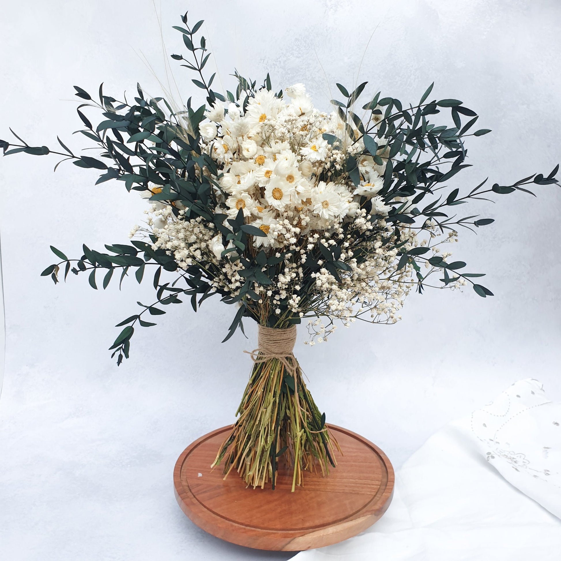 "Snowdrop Glade" Minimalist Large Handtie Bouquet - Lytton Rose Botanical. "Snowdrop Glade" Minimalist Large Handtie Bouquet - Lytton Rose Botanical. A beautifully arranged bouquet of dried flowers. Delicate white daisies are the centerpiece, surrounded by lush green eucalyptus leaves and wispy baby's breath. The bouquet is elegantly tied with twine and displayed on a rustic wooden board.
