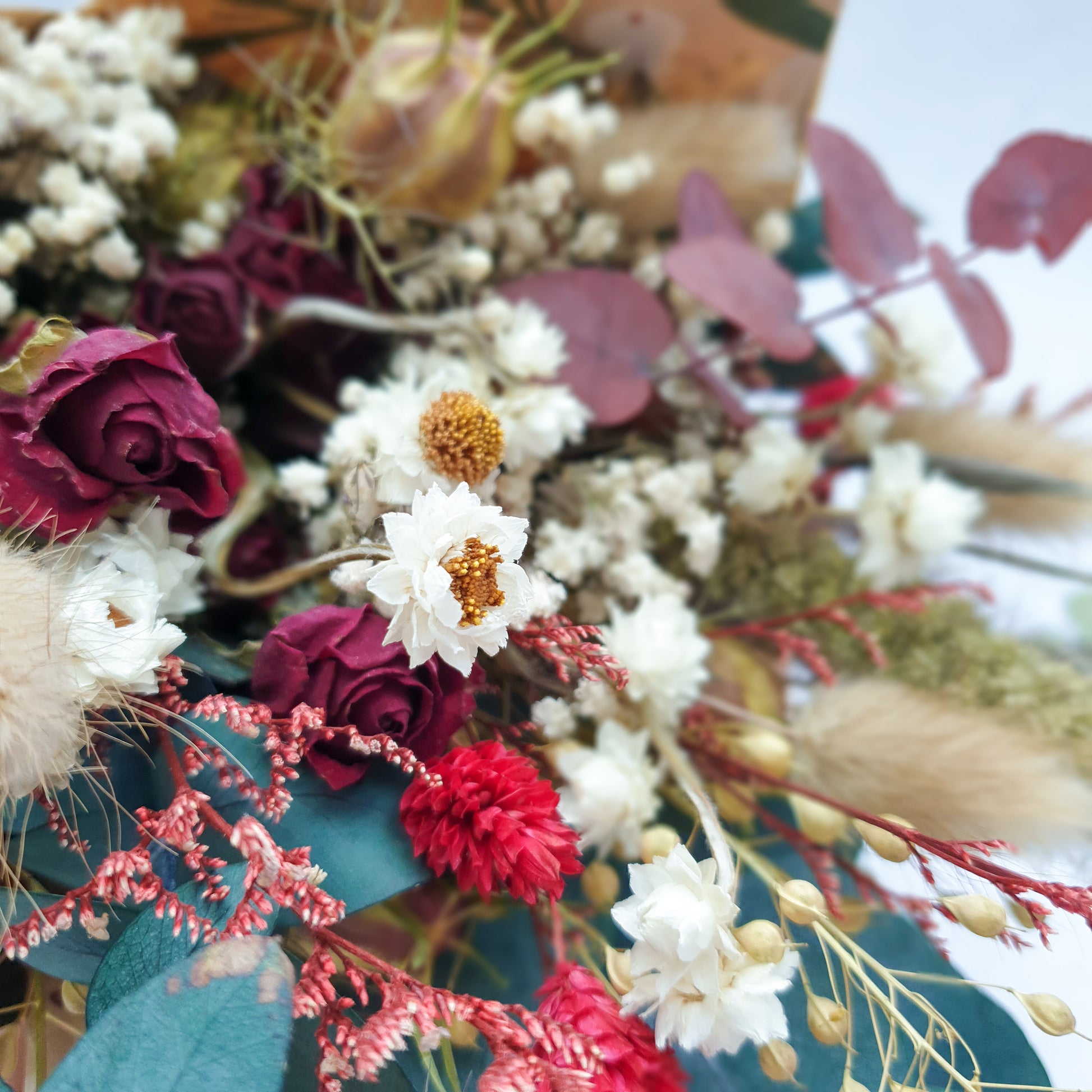"Winter Woodlands" Preserved Eucalyptus & Dried Roses Small Bouquet - Lytton Rose Botanical. A detailed close-up image highlighting the intricate details of a dried flower arrangement. Delicate white flowers with yellow centers, vibrant red roses, and lush green eucalyptus leaves create a stunning contrast.