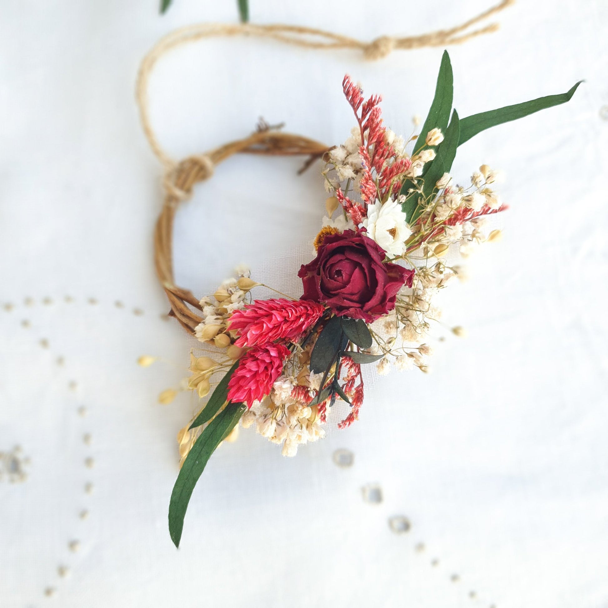 Mini Wreath by Lytton Rose Botanical. A delicate, circular tiny wreath crafted from dried flowers and twigs. The wreath features a cluster of cream-colored dried flowers, including gypsophila and statice, with a single vibrant red rose as the centerpiece. Lush green eucalyptus leaves frame the arrangement. The wreath is secured with a thin, natural-colored twine.
