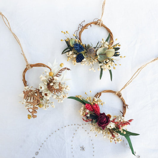 Set of 3 Mini Wreaths by Lytton Rose Botanical. Three delicate, tiny wreaths crafted from dried flowers and twigs. The first wreath features a cluster of white dried flowers, including daisies and gypsophila, interspersed with dried brown ferns. The second wreath showcases a mix of white dried flowers, blue thistle-like flowers, and green eucalyptus leaves. The third wreath features a vibrant red rose as the centerpiece, surrounded by cream-colored dried flowers and red accents. 