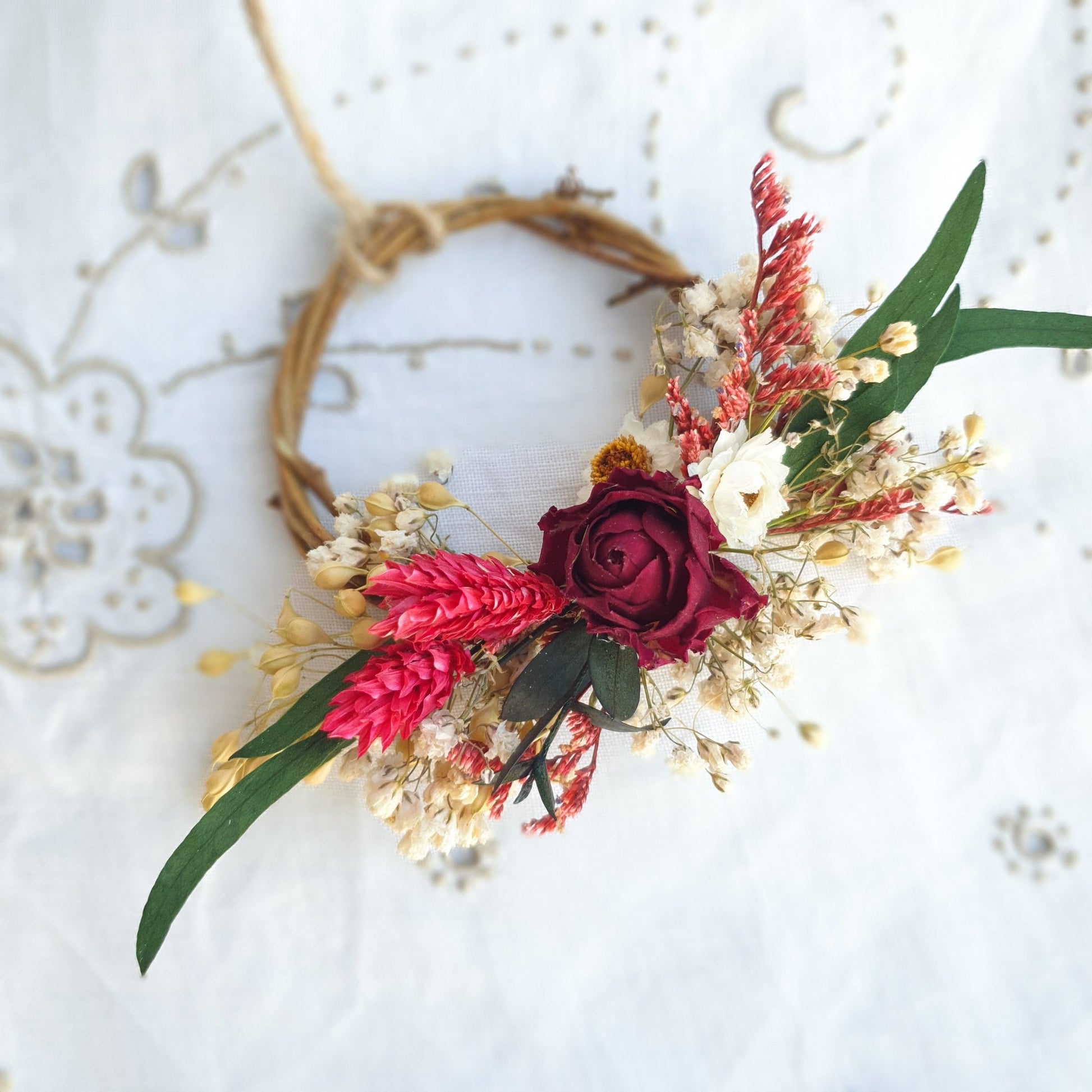 Mini Wreath by Lytton Rose Botanical. A delicate, circular tiny wreath crafted from dried flowers and twigs. The wreath features a cluster of cream-colored dried flowers, including gypsophila and statice, with a single vibrant red rose as the centerpiece. Lush green eucalyptus leaves frame the arrangement. The wreath is secured with a thin, natural-colored twine.