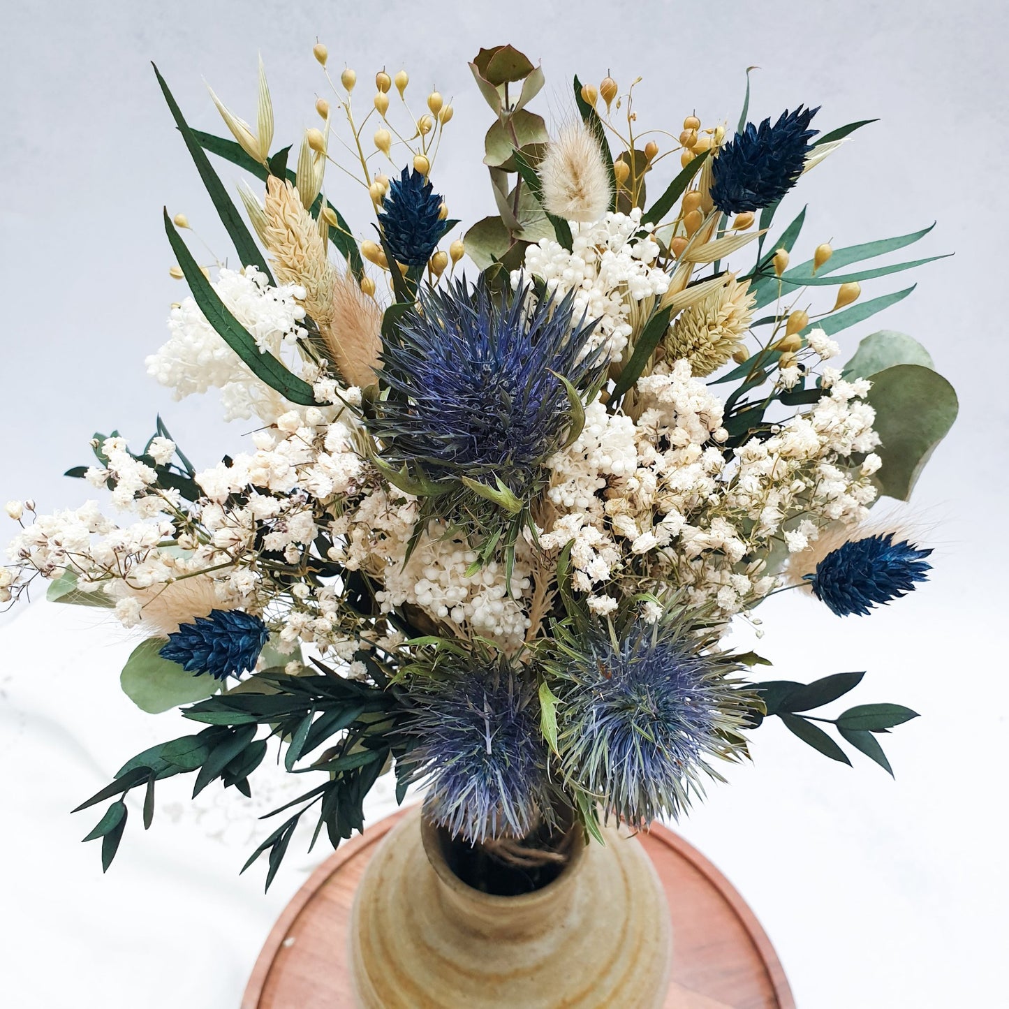 "Frosted Forest" Dried and Preserved Small Eucalyptus Bouquet - Lytton Rose Botanical, A close up of a brown ceramic vase filled with a vibrant bouquet of dried flowers. The bouquet features a mix of white gypsophila, blue thistles, and green eucalyptus leaves. The vase is displayed on a wooden tray.