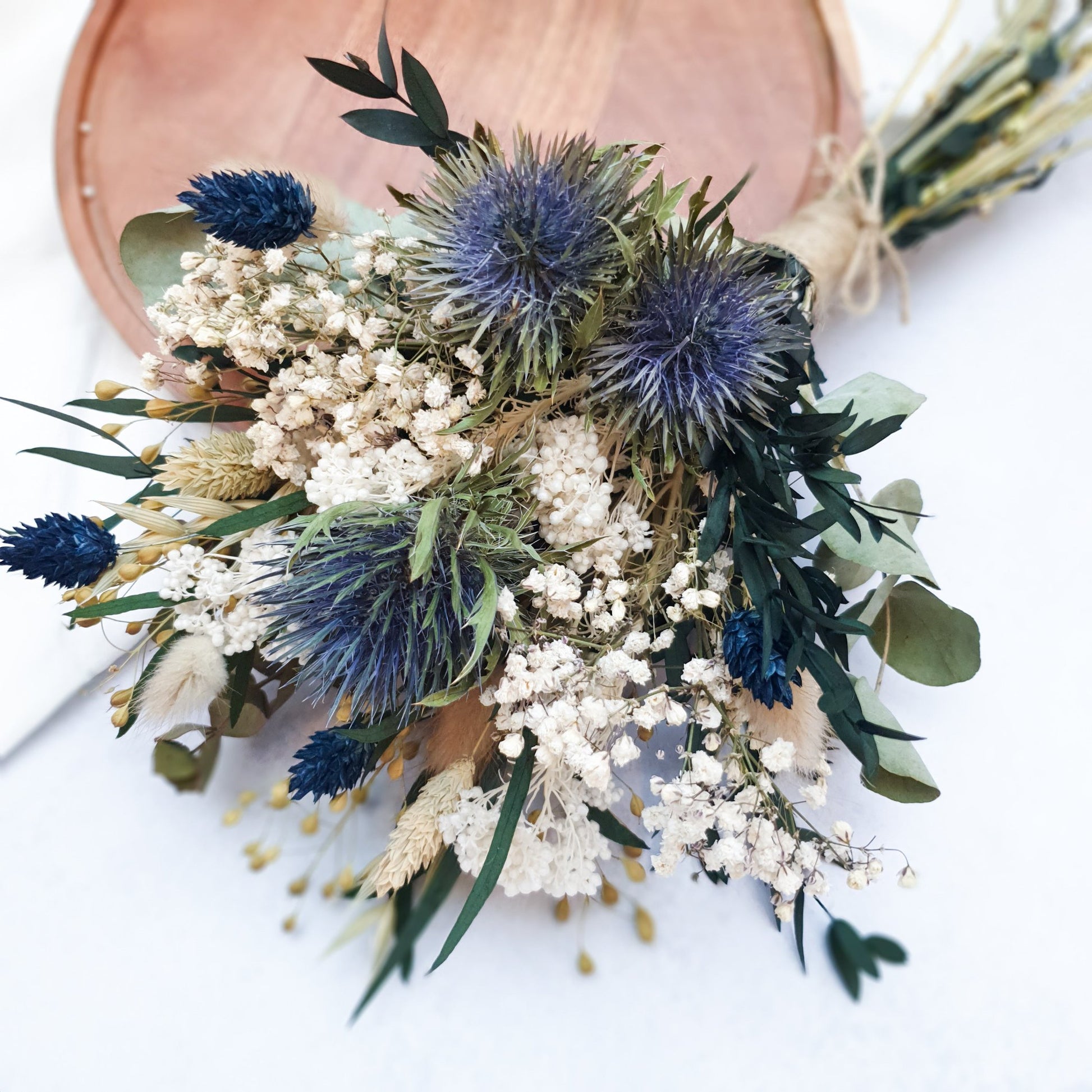 "Frosted Forest" Dried and Preserved Small Eucalyptus Bouquet - Lytton Rose Botanical. A bouquet of dried flowers, primarily white and blue, with green foliage. The bouquet is tied with twine and laid on a rustic brown board.