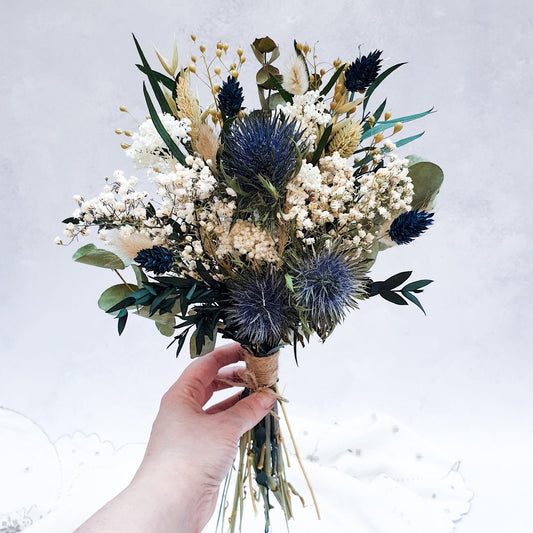"Frosted Forest" Dried and Preserved Small Eucalyptus Bouquet - Lytton Rose Botanical. A hand holding a beautifully arranged bouquet of dried flowers. Delicate white flowers, vibrant blue thistles, and lush green eucalyptus leaves create a stunning contrast. The bouquet is elegantly tied with twine.