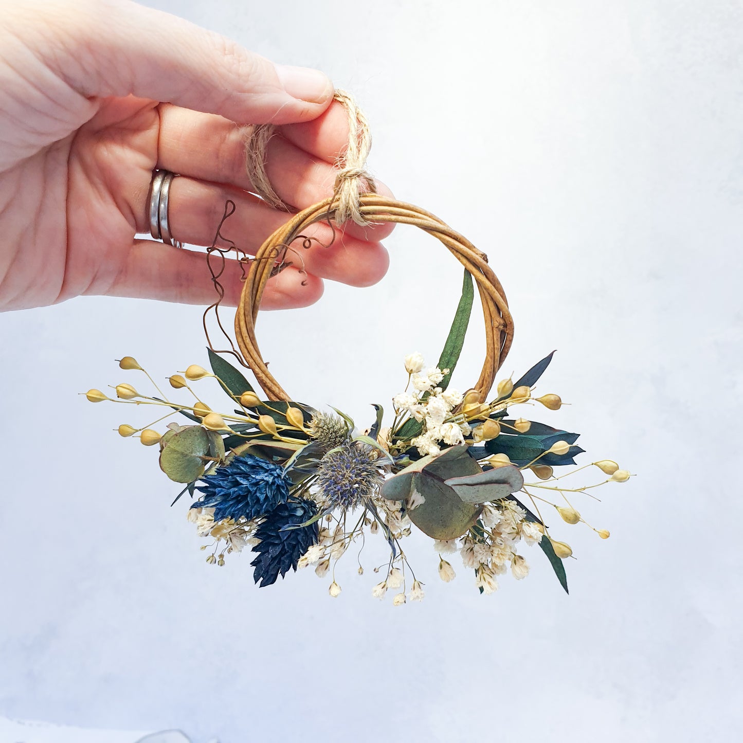 Mini Wreaths - Lytton Rose Botanical. A delicate, circular wreath crafted from dried flowers and grapevine. The wreath features a cluster of blue and cream-colored dried flowers, including gypsophila and canary grass, interspersed with vibrant blue thistle-like flowers and lush green eucalyptus leaves. The wreath is secured with a thin, natural-colored twine.